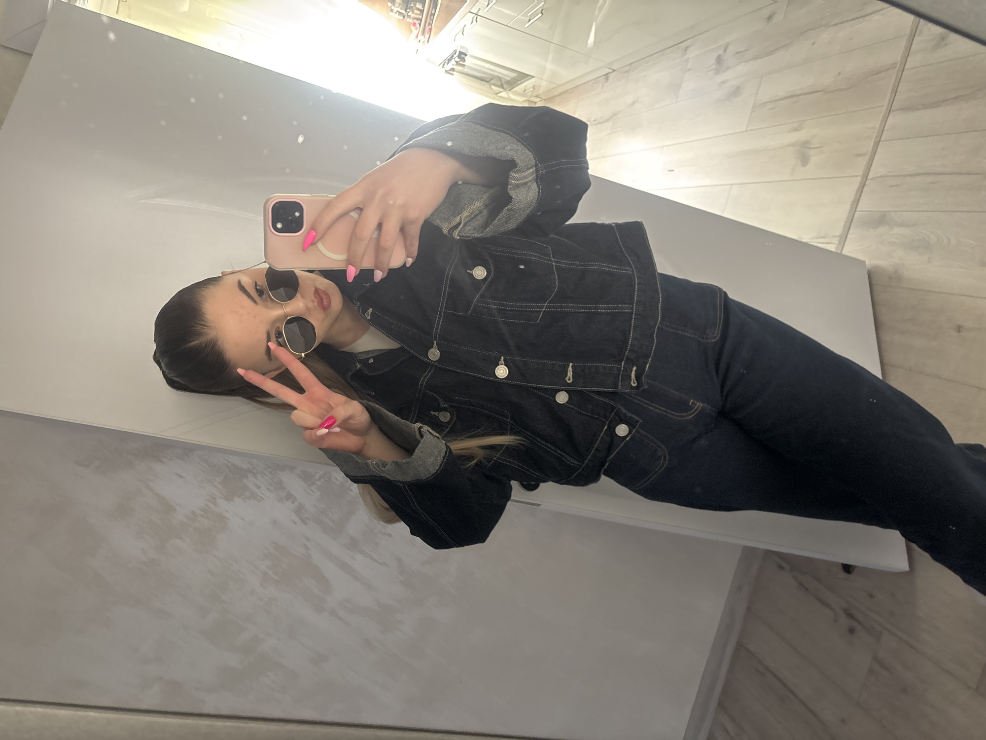 A young woman with dark hair pulled back is taking a selfie in front of a mirror. She is wearing a dark denim jacket with gray lining on the cuffs and a white t-shirt. She also has sunglasses on and pink nail polish. She is holding up a pink phone in her right hand and making a peace sign with her left hand. The background consists of a reflective surface on the left and a tiled floor on the right. There is nothing that definitively identifies the location in Plovdiv, Bulgaria.