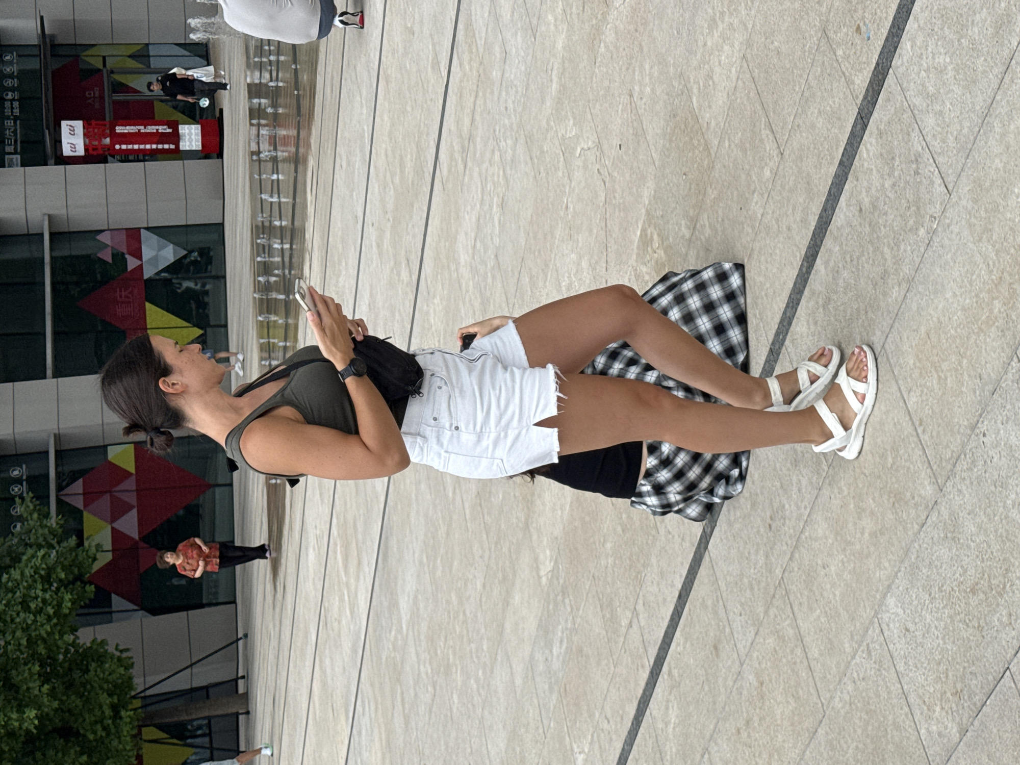A woman in a green tank top and white denim shorts is standing on a paved plaza and looking at her phone. She is wearing white sandals and has a black fanny pack across her chest. Behind her, a person in a black and white plaid skirt is sitting down. In the background, a man in a black shirt and pants walks past a red and white sign with Chinese characters. There are fountains in the middle of the plaza, and buildings with large windows are visible. The scene appears to be in the city of Yuzhong District, China.