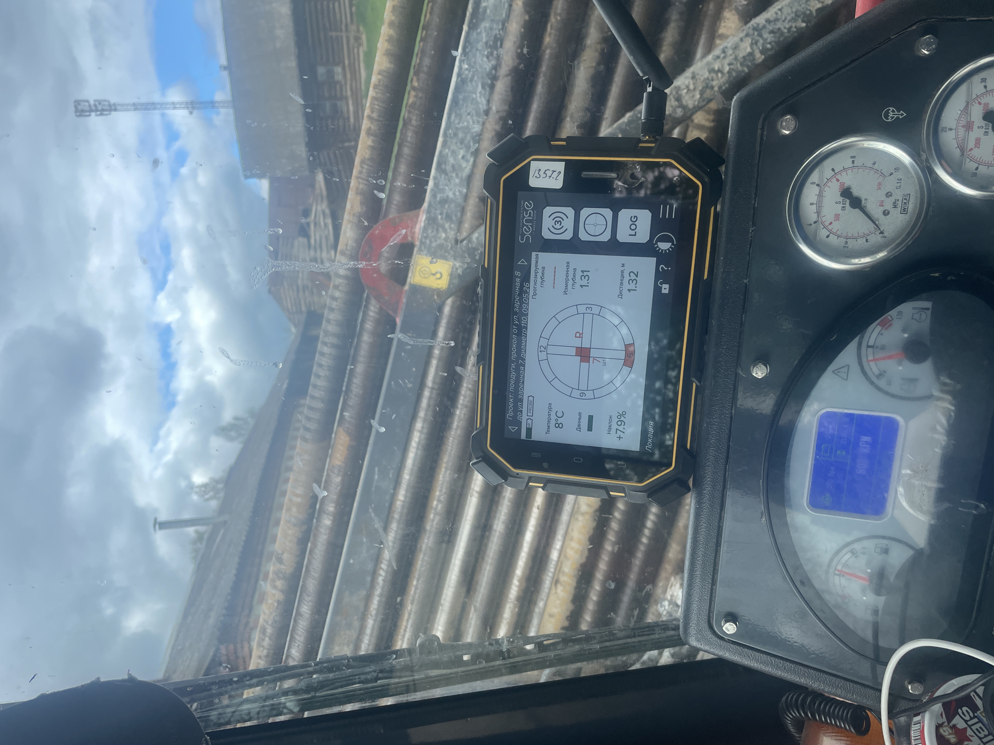 The image shows the interior of a vehicle, presumably a piece of heavy machinery, judging by the dashboard gauges. A smartphone-like device with the brand "Sense" is mounted on the windshield. The device's screen displays data related to a project described as "Проект: поедуги, прокол от ул. заречная 8 до ул. заречная 7, диаметр 110, 09.05.26" (Project: Poedugi, puncture from Zarechnaya St. 8 to Zarechnaya St. 7, diameter 110, 09.05.26). The screen also shows measurements: a predicted depth, current temperature of 8°C, inclination of +7.9%, and a distance of 1.32m.
Through the windshield, a blurred view of pipes or beams and a utility pole is visible. The sky is overcast with a mix of clouds and blue patches. A portion of the dashboard with gauges is visible. The gauges include a pressure gauge with the brand "WIKA" and RPM values. A sticker with the name "Sibis" is on the bottom right.
