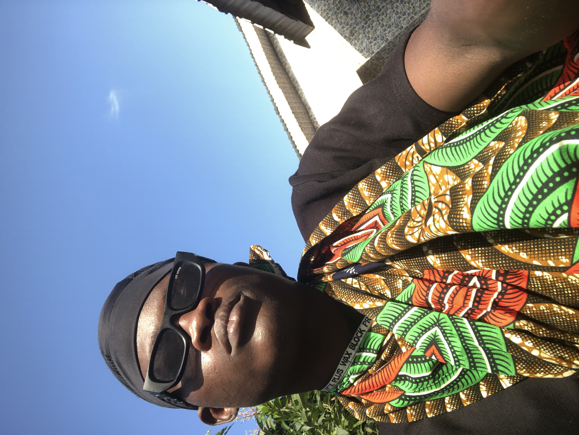 A person is captured in a close-up, low-angle selfie outdoors under a clear blue sky. They are wearing a black durag, black sunglasses with white lettering on the lenses, and a black t-shirt adorned with a vibrantly patterned Ankara fabric wrapped around their neck and chest. The fabric features a bold design of green, red, and brown motifs. The background shows a glimpse of green foliage on the left and a building with a corrugated roof and stone facade on the right. The lighting suggests it is daytime, casting shadows and highlighting the person's facial features. The overall scene conveys a casual, sunny outdoor setting, likely in Ado, Nigeria, given the cultural attire.