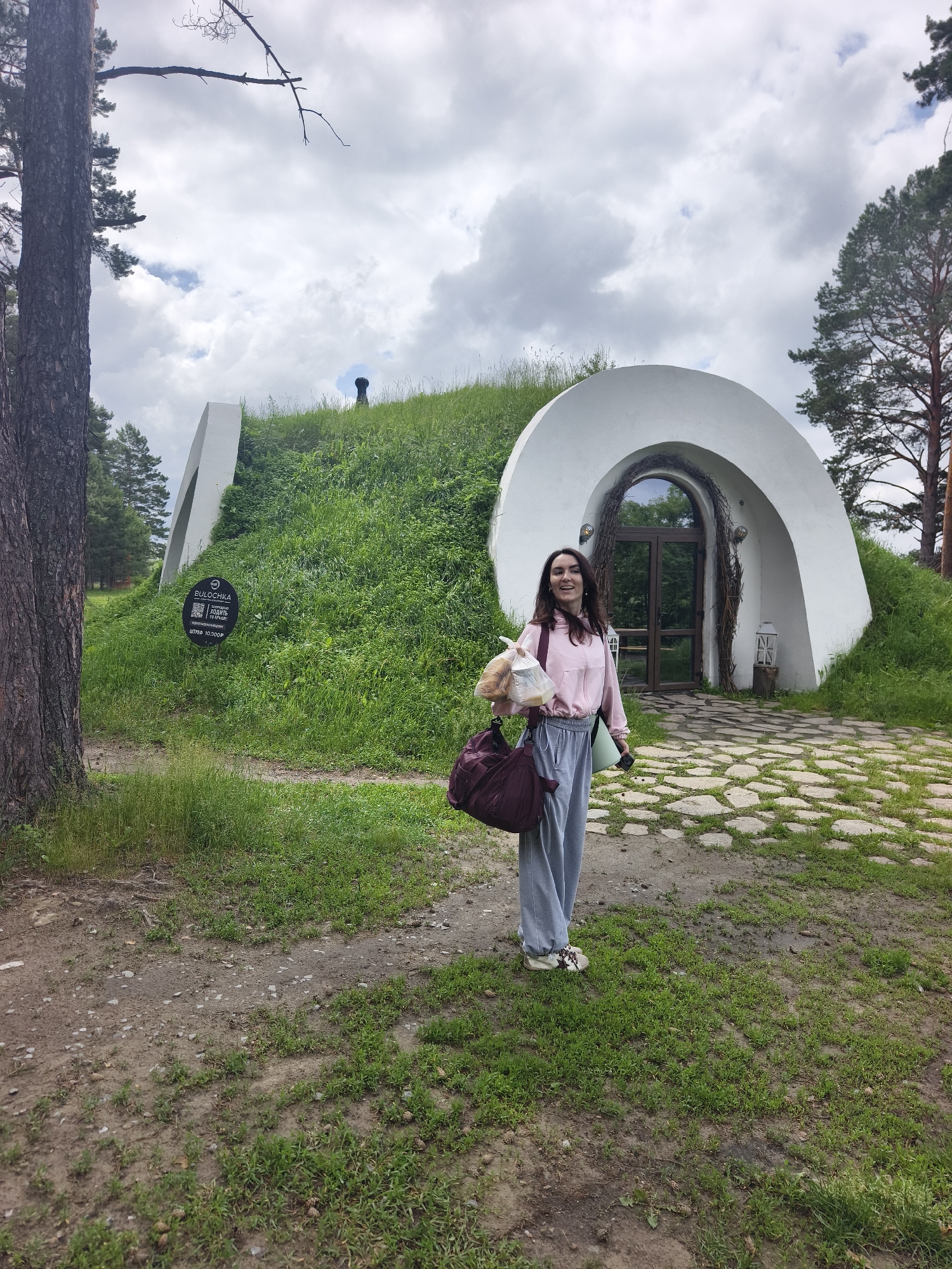 In Онохино, Russia, a woman stands outside a unique, hobbit-hole-like structure, a modern dwelling integrated into a grassy hillside. The building features a rounded white archway framing a wooden door and window, with naturalistic twig decorations. The woman, wearing a pink hooded sweatshirt and grey sweatpants, holds a plastic bag containing food and a maroon duffel bag. Her feet are clad in casual sneakers. A green yoga mat is tucked under her arm. The ground leading to the entrance is paved with irregular stone slabs. A dark sign with white text and a QR code is affixed to the side of the grassy mound to the left of the entrance. Surrounding the structure are lush green grass, trees, and a cloudy sky.