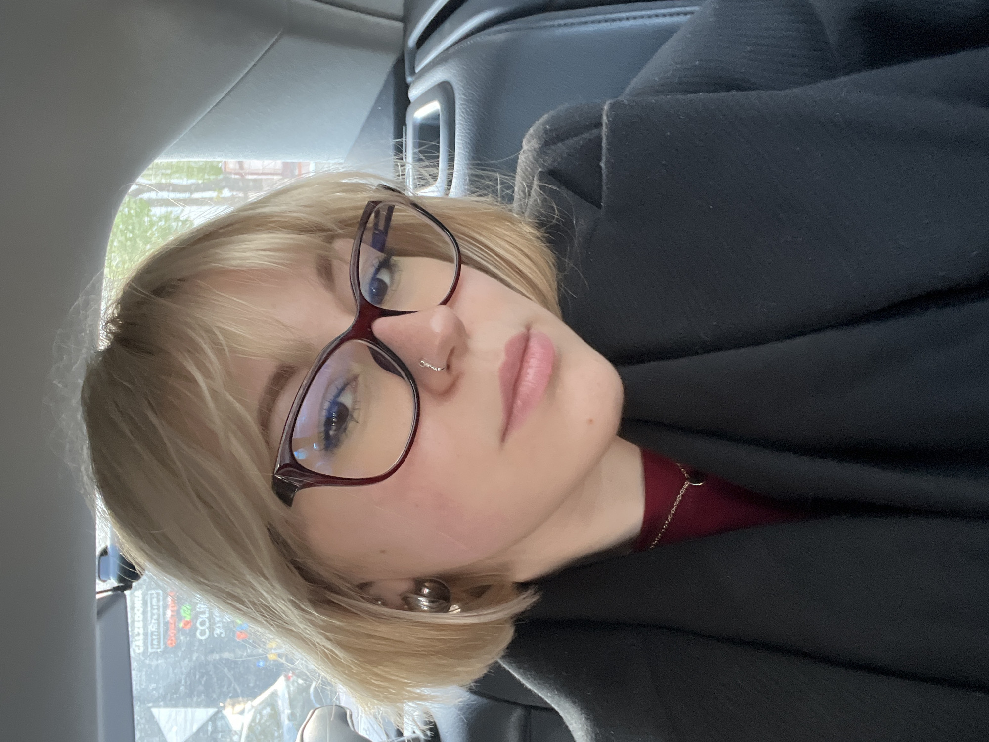 A young individual with short, choppy blonde hair and bangs is pictured in a close-up, head-and-shoulders portrait, taken inside a vehicle. Their head is tilted, and they are looking directly at the camera through dark-rimmed glasses, possibly burgundy or dark red. They have a subtle nose ring (a small silver hoop) and a prominent silver, spherical earring on their left earlobe. Their lips are lightly colored, and light-colored eyeliner is visible above their eyes.
They are wearing a dark, possibly wool or felt, outer garment (like a coat or jacket) over a dark red top or shirt, from which a thin gold chain necklace is visible. The texture of the dark garment is noticeable in several areas.
The setting is the interior of a car, with parts of the grey headliner and dark seats visible in the background. Through the car window on the left, an outdoor scene is visible. This includes some greenery, possibly trees or bushes, and in the reflection or beyond, illuminated advertisements or store signs. Visible text on these signs includes "CALZEDONIA" and a partial "COUP". The natural light suggests it is daytime. The individual's expression is neutral. The presence of commercial signage like "CALZEDONIA" is consistent with an urban environment like Odesa, Ukraine.