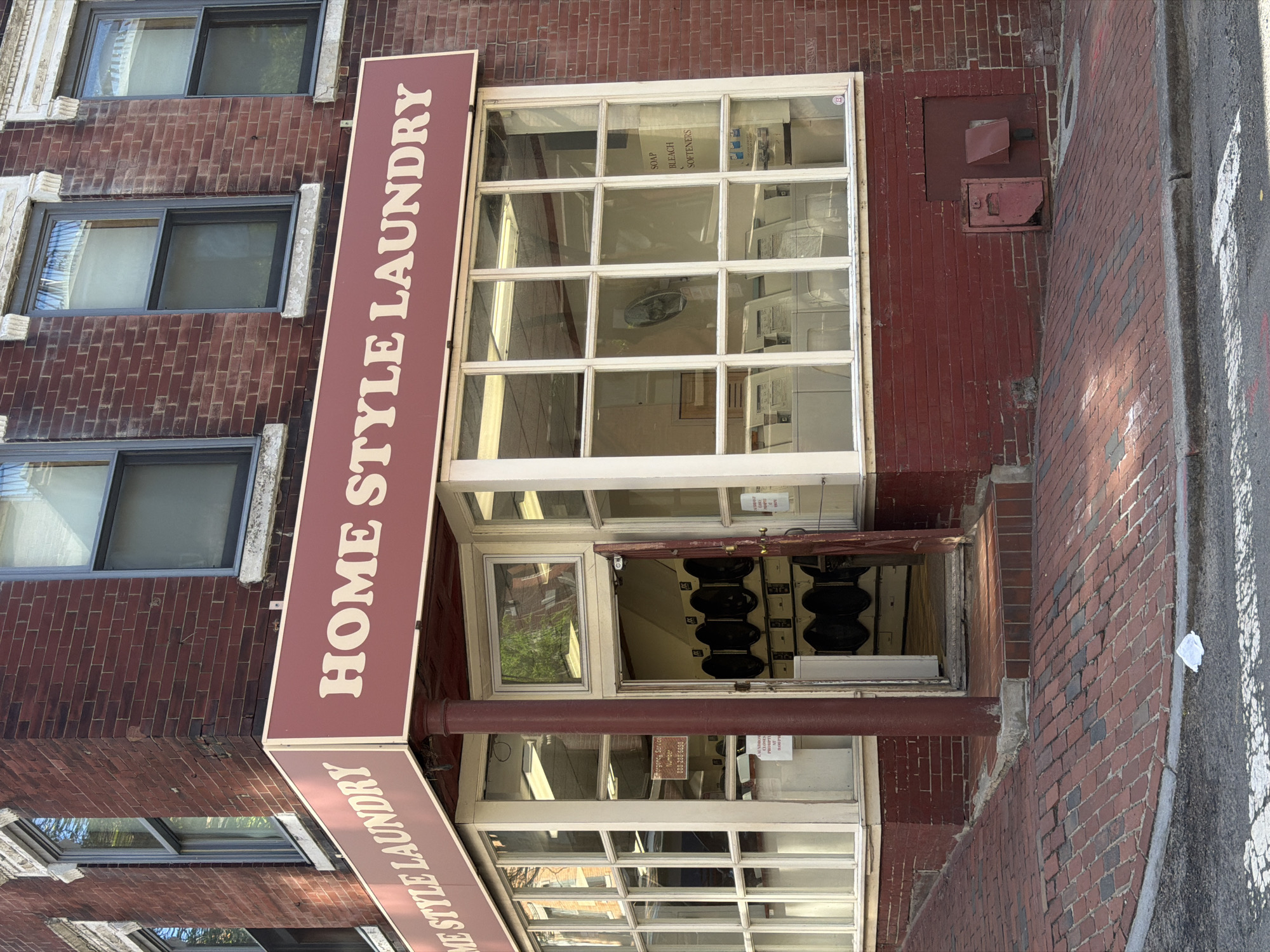 The image shows the facade of a brick building in Boston, Massachusetts, featuring a "Home Style Laundry" storefront. The storefront is characterized by large, multi-paned windows, a brown awning with white lettering reading "HOME STYLE LAUNDRY," and a recessed entrance. The interior of the laundry is partially visible through the windows, revealing rows of washing machines. The building is of older construction, with weathered brick and decorative window frames. Adjacent to the storefront, the brick sidewalk meets the curb of a city street. A metal utility box is visible on the wall next to the laundry.
