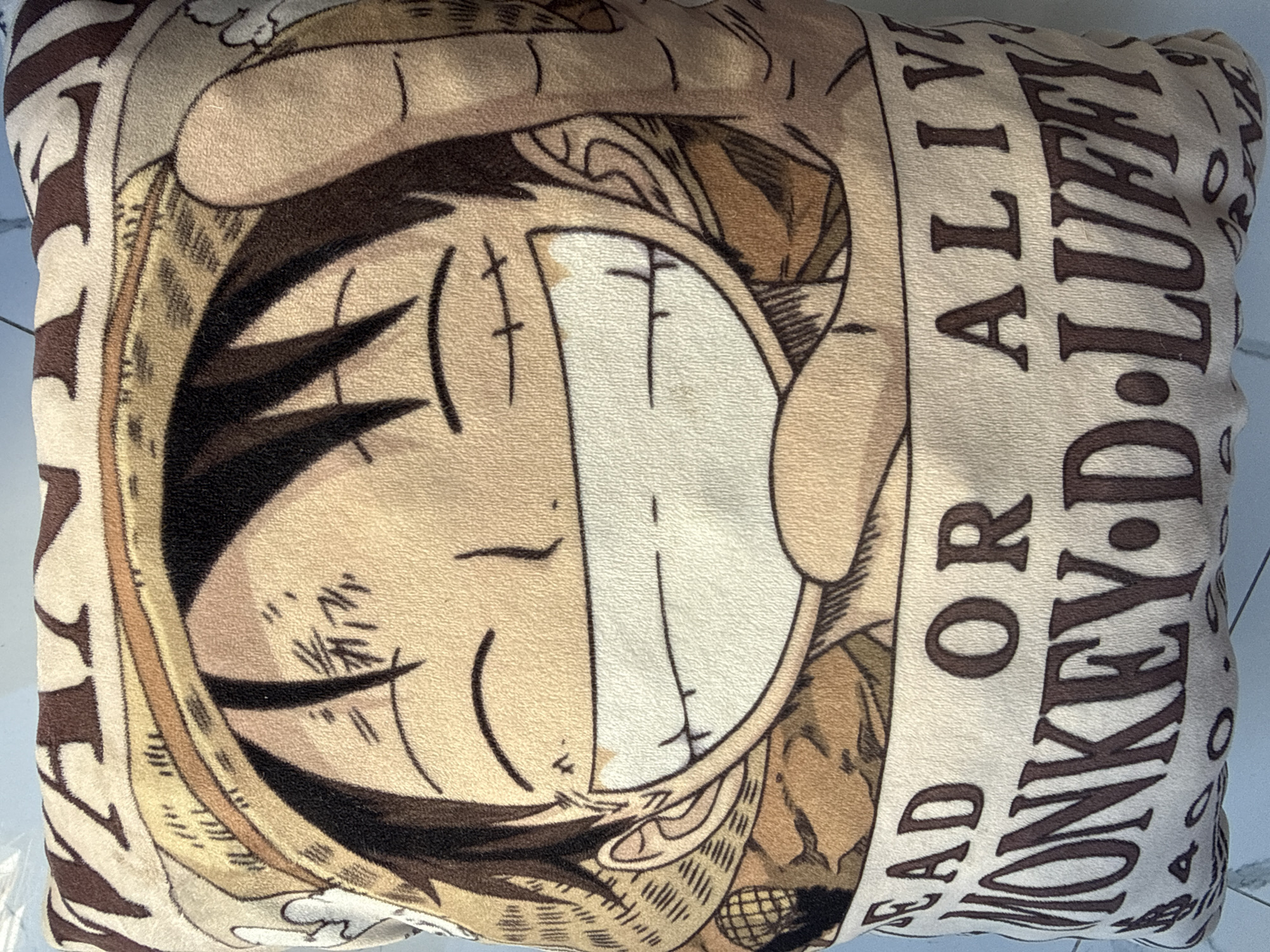 The image is a close-up, overhead shot of a plush pillow featuring a character from the anime series One Piece. The character, identified by the text on the pillow as "MONKEY D. LUFFY," is depicted with a wide grin and wearing his signature straw hat. The pillow has a textured, fuzzy surface. The text on the pillow reads "DEAD OR ALIVE" and "MONKEY D. LUFFY." The background of the pillow has a muted, sepia-toned pattern. The pillow is situated on a light-colored, flat surface that appears to be a floor or table. The lighting is natural and casts soft shadows. There is no indication of the city of Bac Nha Trang Ward, Vietnam, in this image.