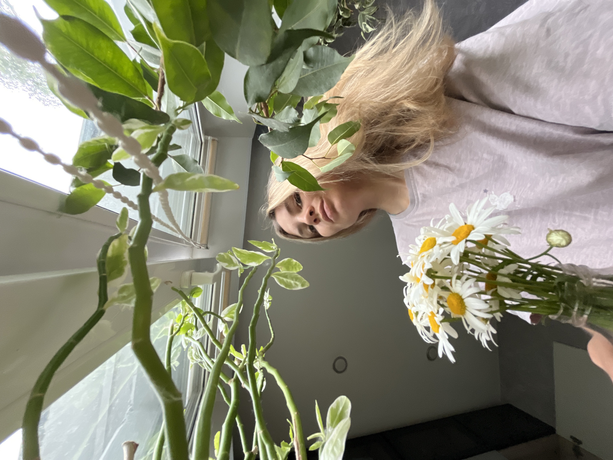 A young woman with long blonde hair, wearing a pale purple t-shirt, is holding a bouquet of daisies in a glass vase. She is standing in front of a large window or skylight, with green plants hanging in the foreground. The background is a room with grey walls and a white ceiling. The image appears to be taken indoors, possibly in a home or a greenhouse. There is no discernible indication of the location being in Chaikovsky, Russia.