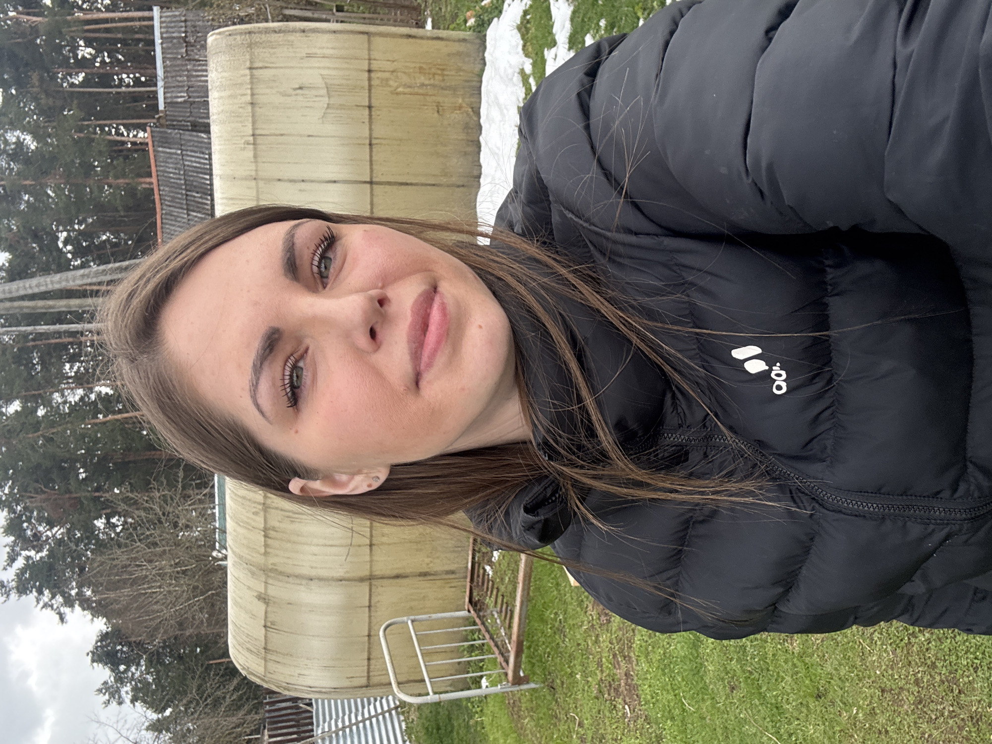 A young woman is captured in a selfie-style portrait taken outdoors in Razdolye, Russia, under daylight conditions with an overcast sky. She has long, dark brown hair, light-colored eyes (appearing green), and a subtle smile directed at the camera. She is wearing a black puffer jacket, on which a partial white logo "o!" (part of the Adidas brand) is visible on the chest.
The background suggests a rural or semi-rural setting. To the left, tall pine trees are prominent, indicating a wooded area. To the right, a large, light-colored, possibly cylindrical or arched structure, resembling a tank or part of a shed, stands. Behind it, a building with a dark, corrugated roof can be seen. The ground is covered with green grass interspersed with patches of melting snow, suggesting a late winter or early spring season. A simple metal fence or gate is visible in the lower right foreground. The woman appears relaxed, posing casually for the picture.
