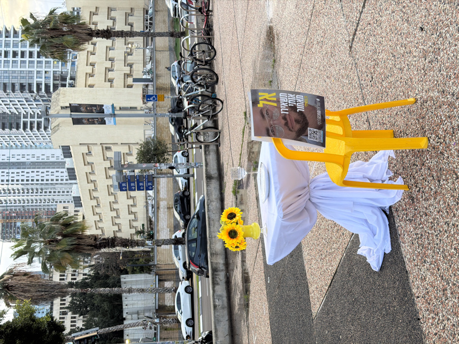 The image captures an outdoor symbolic display situated on a paved pedestrian area in Tel-Aviv, Israel, adjacent to a multi-lane urban street. The background architecture, featuring a large tan building with a distinctive geometric facade and a dense cluster of modern high-rise towers, indicates a location near the Dizengoff Center in central Tel-Aviv.
In the foreground, a small square table, draped with a white tablecloth gathered at its base, holds a yellow vase containing three sunflowers, a white dinner plate, and an empty clear wine glass. Next to the table, a bright yellow plastic chair displays a rectangular poster on its back. The poster features a headshot of a young man with glasses and includes Hebrew text, notably "אל תשאירו אותי מאחור" (Don't leave me behind), along with a QR code and a hashtag, signifying a campaign for a missing individual.
Behind this display, a concrete median separates the pedestrian zone from a street with several vehicles, including white sedans and a black hatchback. A bicycle rack with multiple bicycles is visible further down the street. Utility poles with blue directional signs and a disabled parking symbol stand nearby. Tall palm trees line the street. The prominent tan building in the background exhibits a repetitive pattern of recessed rectangular elements on its facade, characteristic of the Dizengoff Center. Numerous other high-rise residential and commercial buildings populate the skyline. The ground is paved with sections of dark and light grey aggregate, showing evidence of recent moisture. No people are directly interacting with the foreground objects.
