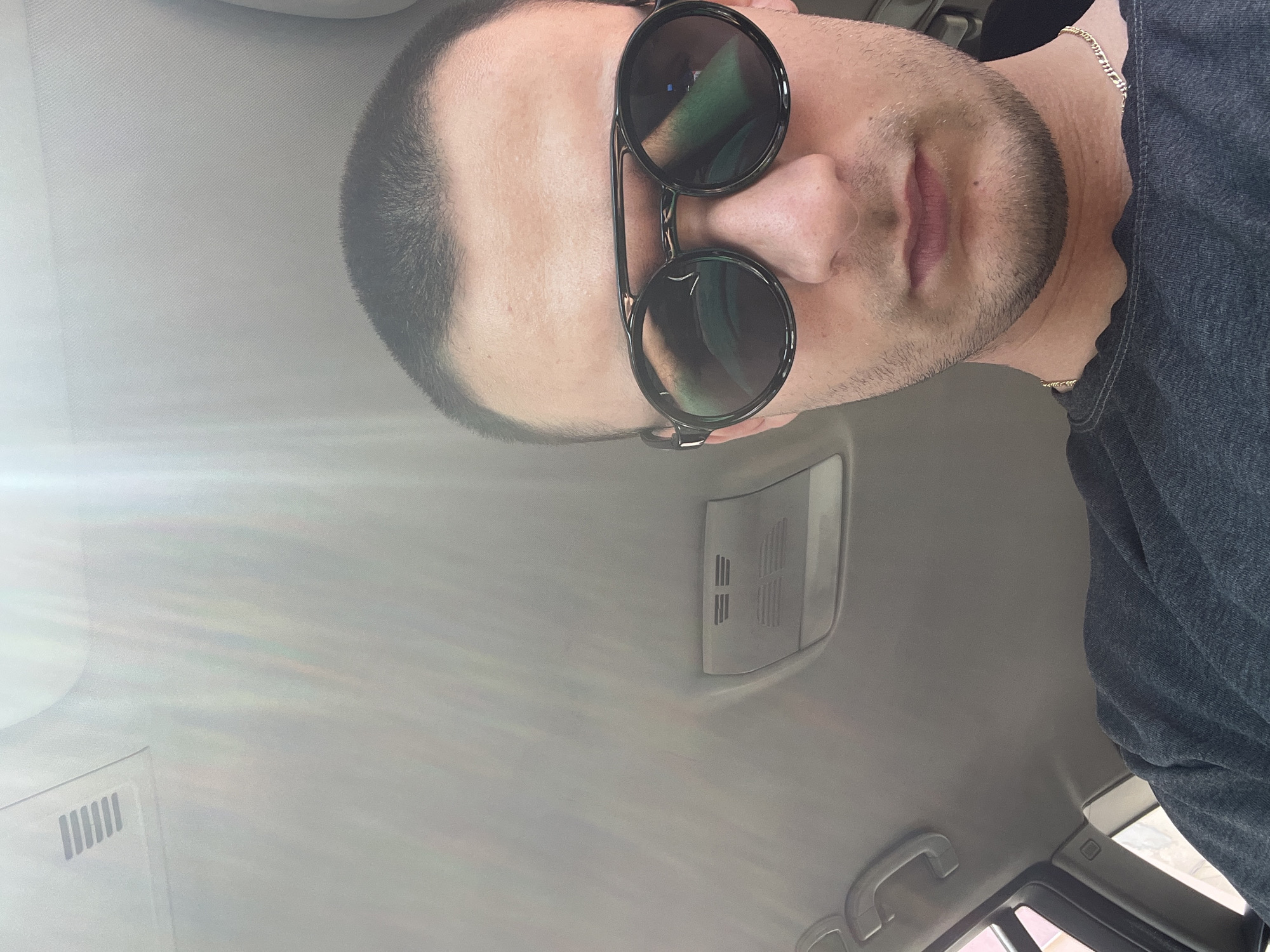 A man wearing round sunglasses and a dark grey shirt is taking a selfie inside a vehicle. The interior ceiling of the car is light grey. The location, Troyan, Bulgaria, cannot be confirmed from the image.