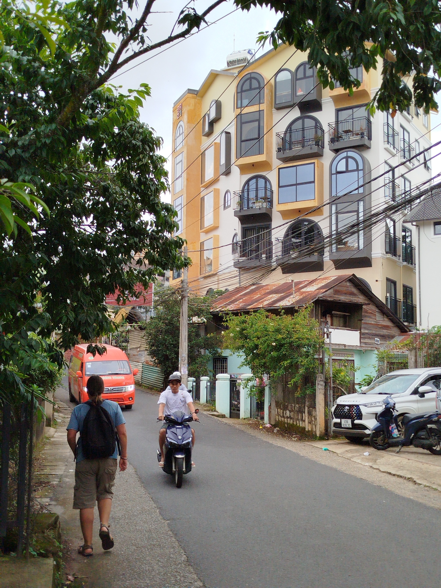 The image depicts a street scene in Da Lat, Vietnam, with a focus on a modern, multi-story building with yellow and white facades and numerous arched windows and balconies. In the foreground, a person with a backpack walks away from the camera on a sidewalk. To the right, on the road, a person on a dark-colored scooter rides towards the viewer. Further down the street, an orange van is visible, and a white car is parked on the right side. The environment includes trees, power lines, and a mix of older and newer structures, suggesting a typical urban landscape in Da Lat.