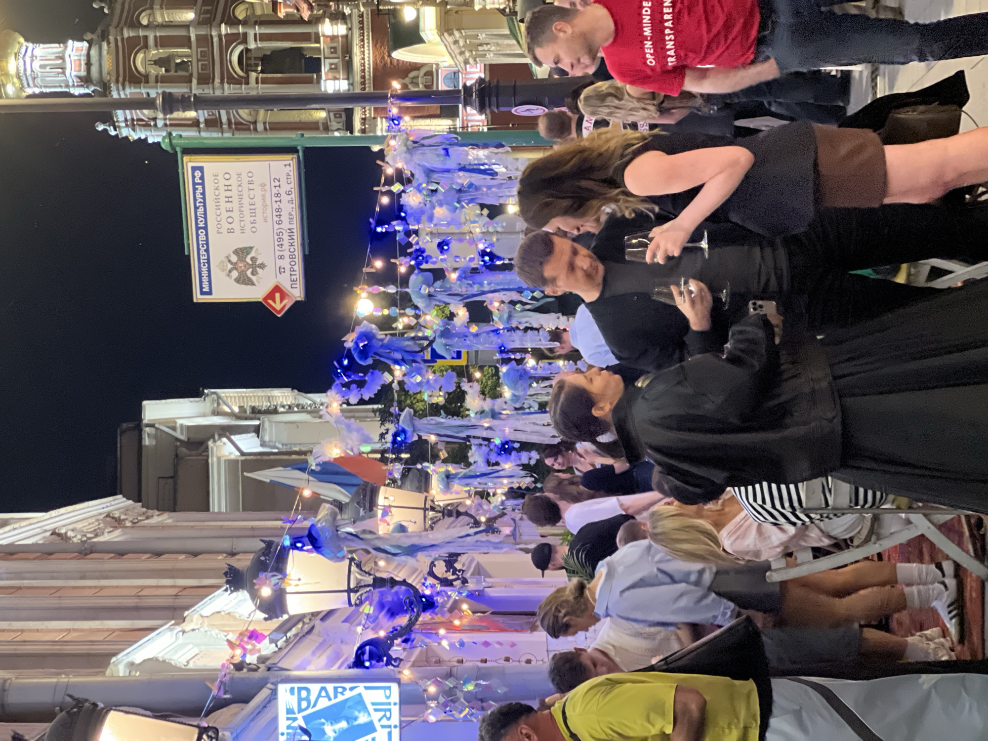 This image depicts a crowded nighttime street scene on Petrovsky Lane, Moscow, Russia. The specific address, "Petrovsky Lane, building 6, structure 1," is visible on a sign for the Ministry of Culture of the Russian Federation and the Russian Military Historical Society.
The street is illuminated by various light sources, including traditional street lamps, decorative string lights with colorful and blue/white bulbs, and a blue neon "BAR" sign on a building to the left. Numerous translucent, blue and white, abstract fabric or plastic decorations resembling jellyfish are suspended across the street, contributing to a festive atmosphere. Several Russian flags are also visible among these decorations.
A diverse group of adults, predominantly casually dressed, occupies the foreground and midground. In the center, a woman with dark wavy hair wearing a black sleeveless top and a brown skirt holds a wine glass while conversing with a man in a dark long-sleeved shirt who also holds a wine glass. To their immediate left, another woman in a black jacket and long black skirt is partially visible. To the right of the central couple, a man in a red t-shirt bearing the partially visible text "OPEN-MINDED TRANSPAREN" stands with his hands in his pockets. Other individuals are seen standing, walking, or seated at small tables, engaging in conversations and socializing. One woman with blonde hair in a striped shirt is seated at a white foldable chair.
Architecturally, buildings with classical details and illuminated windows line the street. In the upper right background, a distinctive building with a red-brick lower section, ornate white trim, a bell tower, and a golden onion dome is visible. The ground in parts of the foreground appears to be covered by a red patterned rug or mat.