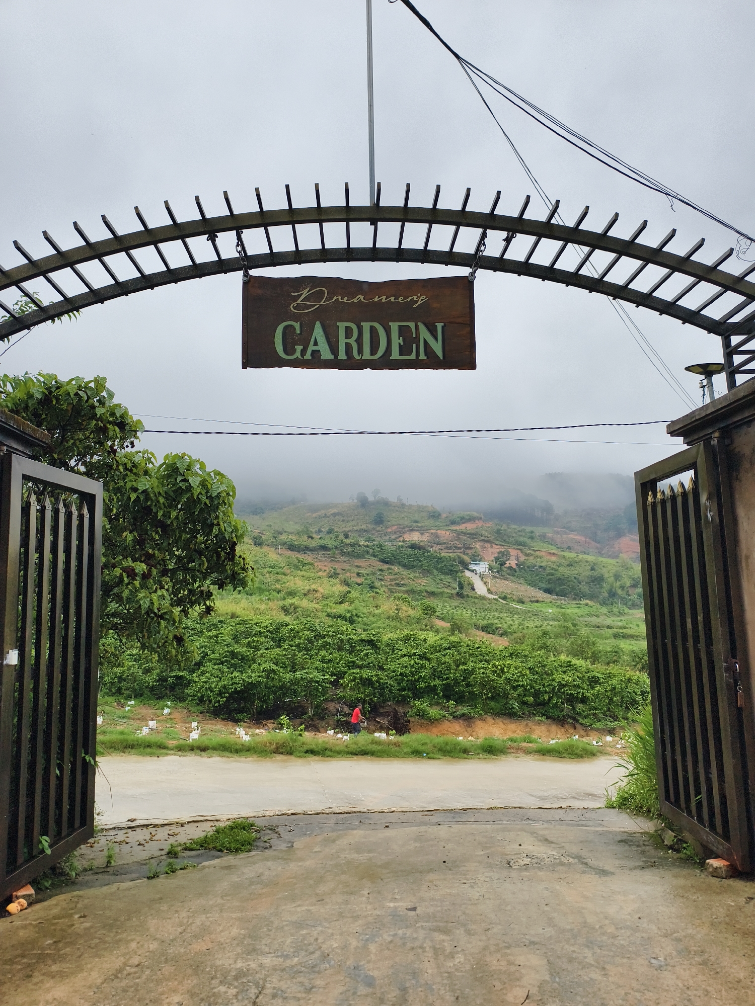 The image shows an open gate leading into "Dreamer's Garden" in Tầm Xá, Vietnam. The gate is under an arched wooden structure with the garden's sign hanging from it. The background features a hillside covered in lush greenery, partially obscured by mist. A small, paved road leads into the garden. A person in a red shirt is visible in the distance, along the road, next to a group of small, white markers or posts. The sky is overcast, creating a muted light.
