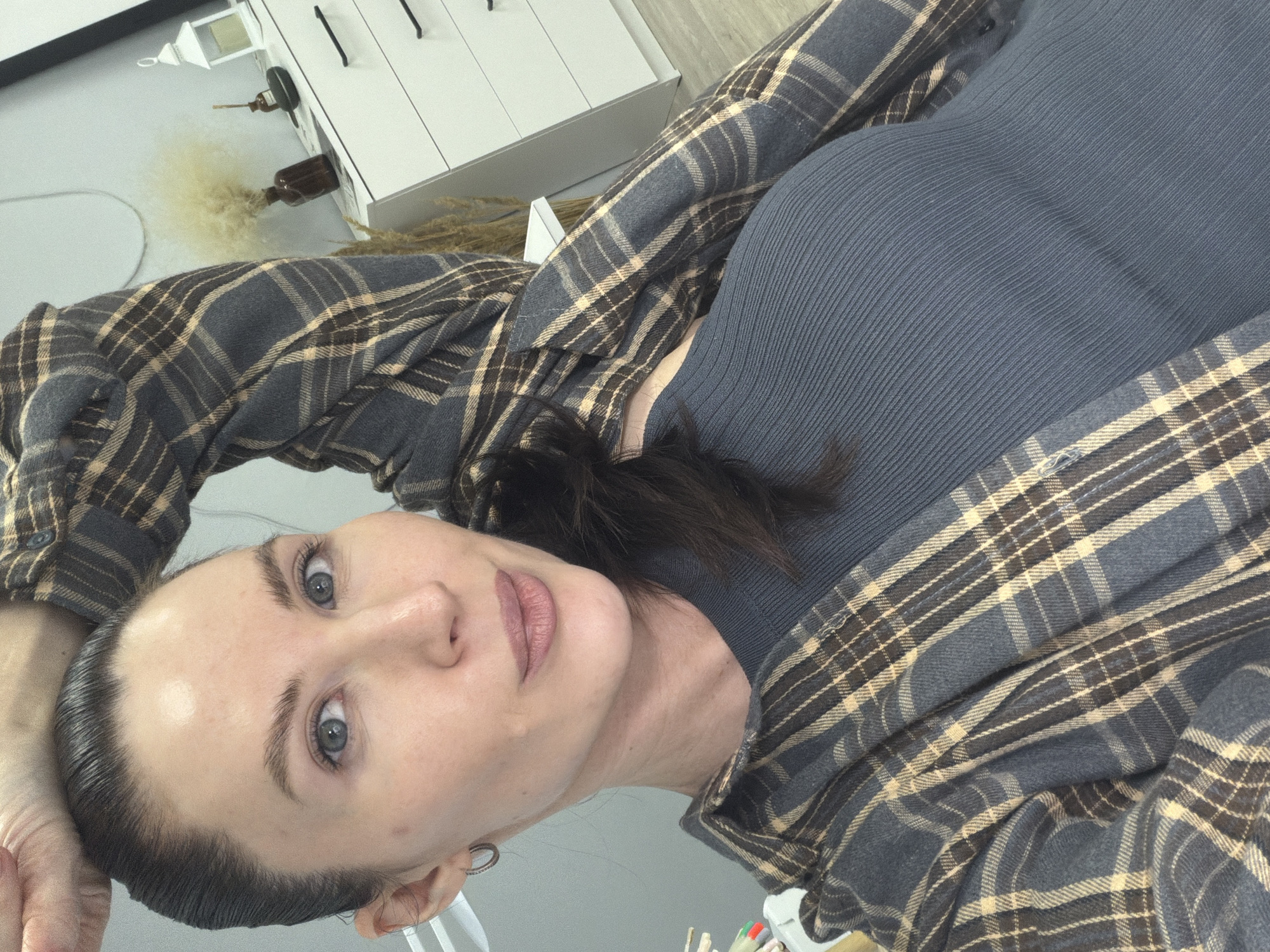 A woman with fair skin and dark hair is facing the camera with a slightly tilted head. She has blue eyes and neutral-toned lips. She is wearing a ribbed, dark gray top underneath a gray and beige plaid flannel shirt. Her left hand is raised to her head. In the background, there is a white cabinet with black drawer pulls, a small white lantern, and decorative grasses. The setting appears to be indoors. No information confirms that the image was taken in Perm, Russia.