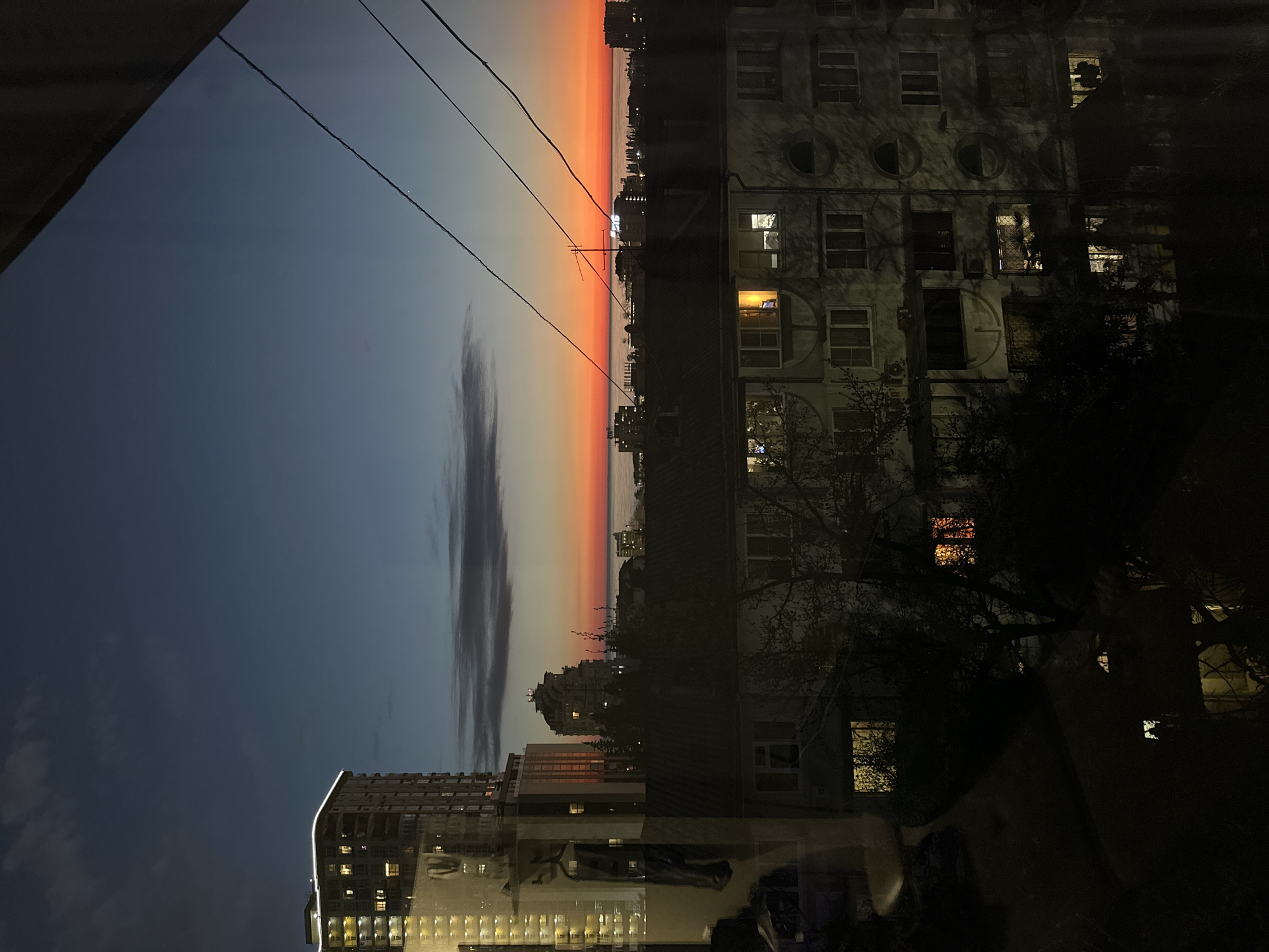 The image shows an evening view of a city with a vibrant sunset. The sky transitions from dark blue to orange, with a dark cloud formation present. High-rise buildings, including one modern structure with many illuminated windows, are visible. Other buildings, older in appearance, have a mix of lit and dark windows. Bare tree branches are also visible. Electrical wires are present in the sky. The scene is likely a view from a window, as reflections are visible. It’s unclear if the location is specifically Sochi, Russia, as there are no definitive landmarks.