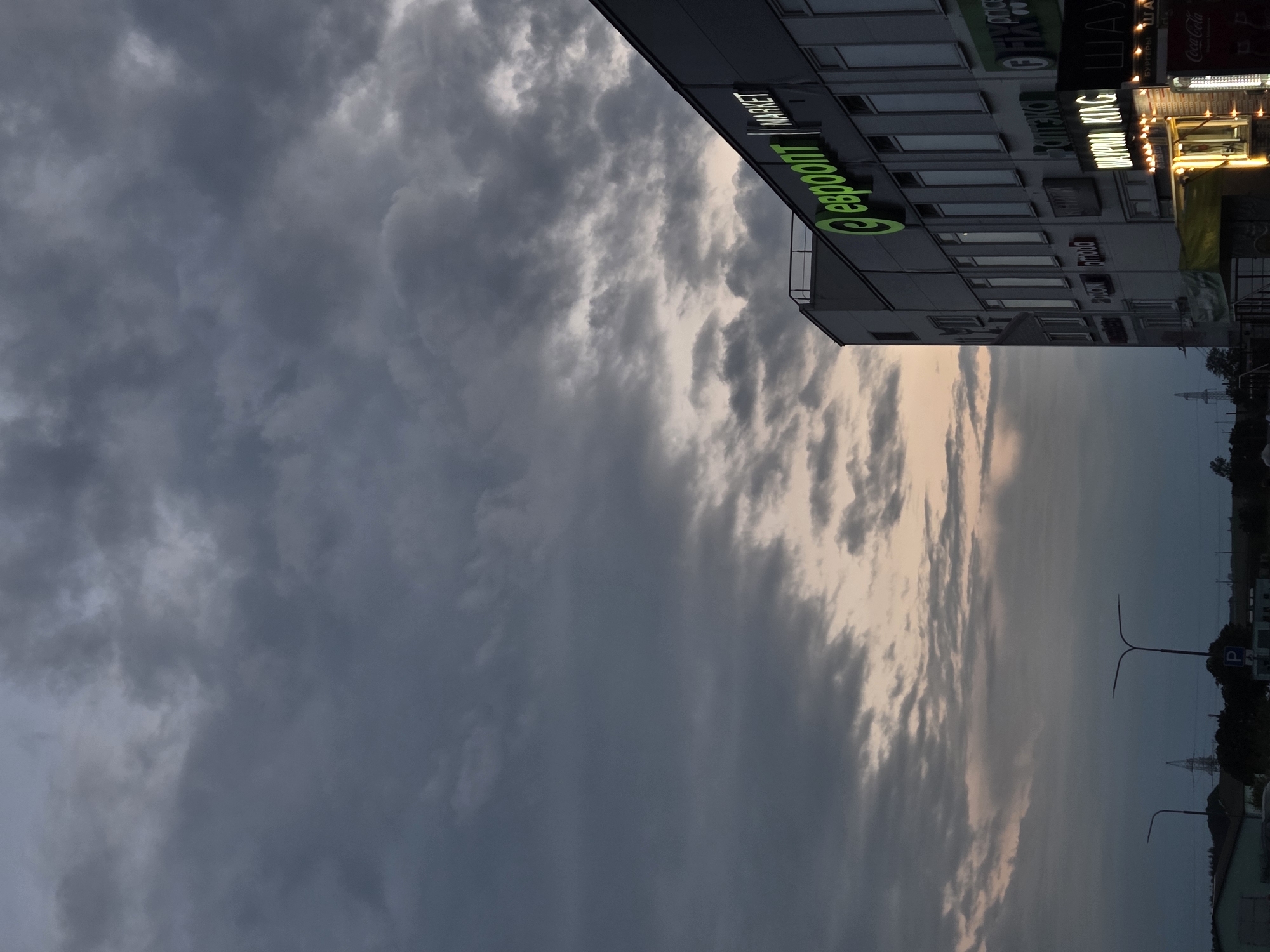 The image captures a cloudy sky over a commercial building in Minsk, Belarus, during what appears to be dusk. The building, with its dark gray facade, features multiple illuminated signs, most prominently a green neon sign that reads "евроопт MARKET" (Evroopt Market). Below this, various other signs are visible, including "Аптека" (Pharmacy), "FIX PRICE," "BURGER KING," and "Coca-Cola." Several windows line the upper stories of the building, and decorative string lights illuminate the lower storefronts. In the background, streetlights and a distant electrical tower are visible, along with silhouetted trees. The overall atmosphere is dim and overcast, with the soft light of the signs contrasting with the darkening sky.