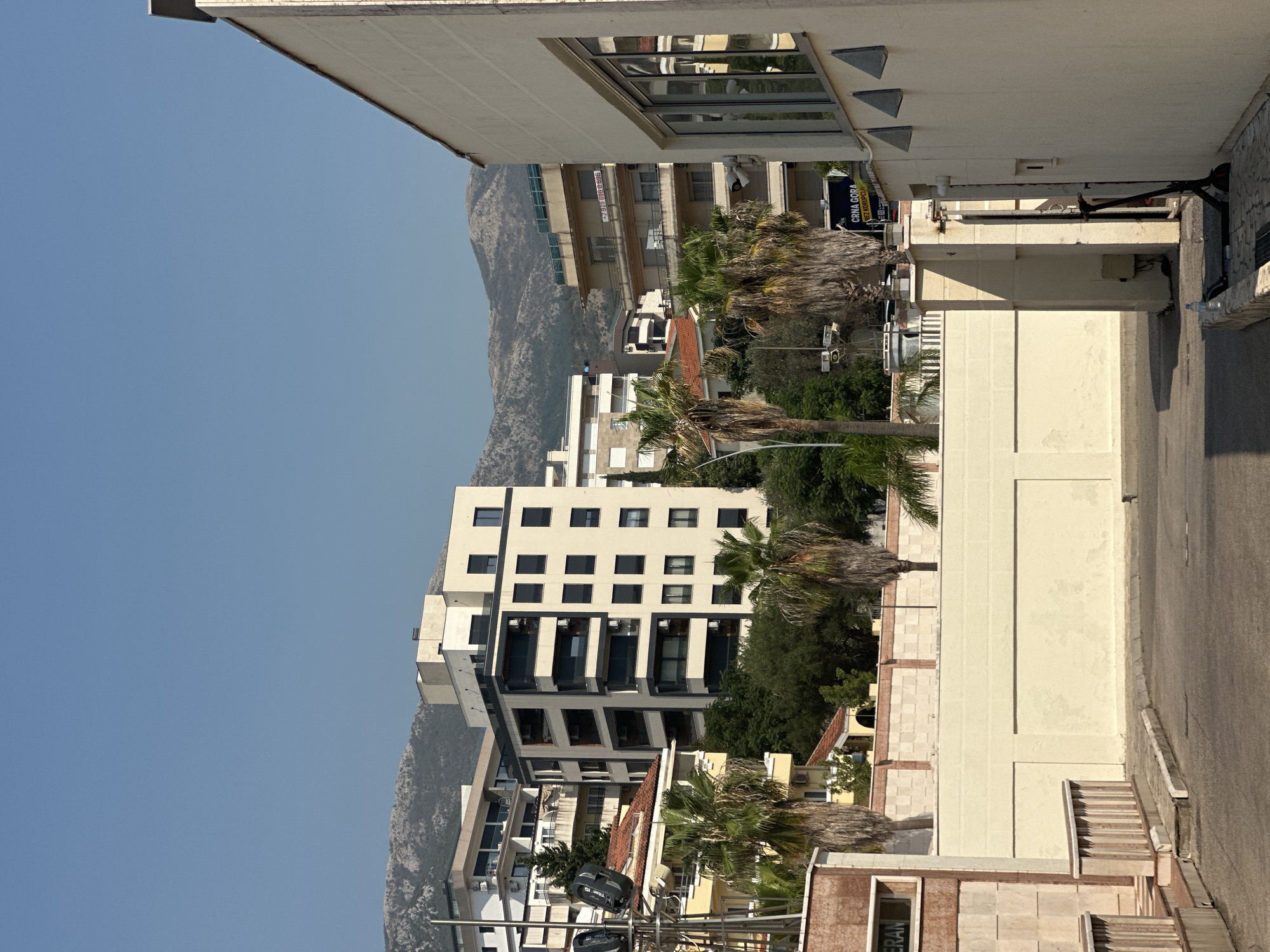 The image depicts a street scene in Boreti, Montenegro. In the foreground, a paved area leads towards a cream-colored building on the right. This building features large windows and triangular decorative elements near the roofline. To the left, a beige wall with a patterned section runs across the middle ground, with a paved walkway and railing in front of it. Beyond the wall, several multi-story apartment buildings are visible, characterized by modern architecture with balconies and numerous windows. Palm trees are scattered throughout the scene, adding a tropical element to the urban environment. In the background, a range of mountains under a clear blue sky completes the vista. A black electric scooter is leaning against the cream-colored building on the right. No people are visible in the image.