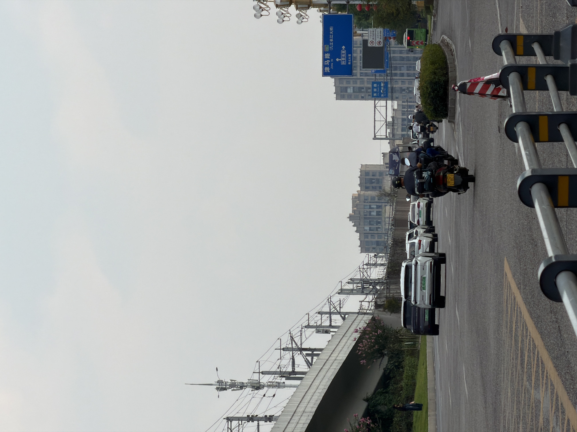 The image depicts a street scene in Jiangjin, China, likely in an urban or suburban area. The foreground shows a paved road with yellow lane markings and a metal barrier. Several vehicles, including cars and motorcycles, are traveling on the road. The cars appear to be modern, with at least one white SUV clearly visible. A few motorcycles are also present, with riders wearing helmets. In the middle ground, there are buildings and infrastructure, including what appears to be an elevated railway or highway with supporting structures and overhead wires. A blue street sign with Chinese characters is visible, indicating directions and road names. In the background, more buildings are present, suggesting a developed urban environment. The sky is overcast with a light haze. Greenery, including trees and bushes, is visible along the side of the road and under the elevated structure. A single person is standing on the grass verge in the left side of the image, looking at their phone.