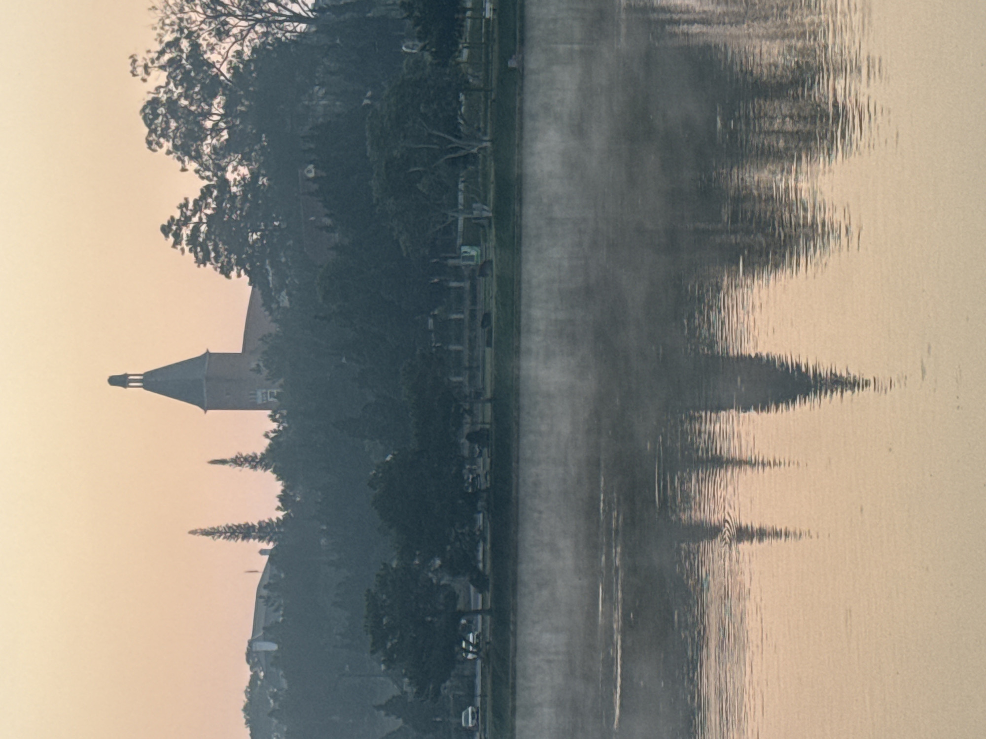 The image captures a misty scene on Xuan Huong Lake in Da Lat, Vietnam. The lake's calm surface reflects the blurred silhouettes of buildings, trees, and a promenade along the shoreline. A prominent structure with a pointed tower stands out among the buildings. The atmospheric conditions create a hazy, muted color palette, with the sky and water blending into a soft, diffused light. The image conveys a sense of tranquility and serenity.