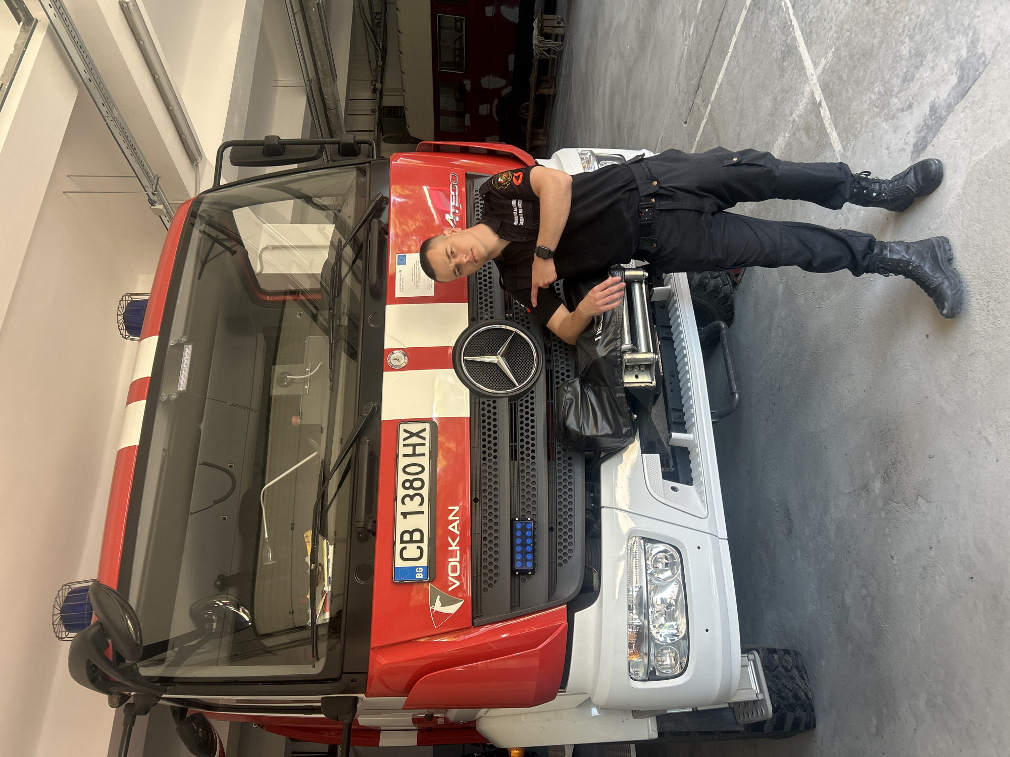 The image shows a fireman in black uniform lying on the hood of a red and white fire truck. The fire truck has a Bulgarian license plate CB 1380 HX, which indicates that the truck is located in Sofia, Bulgaria. The truck is inside a garage, judging by the concrete floor and ceiling.