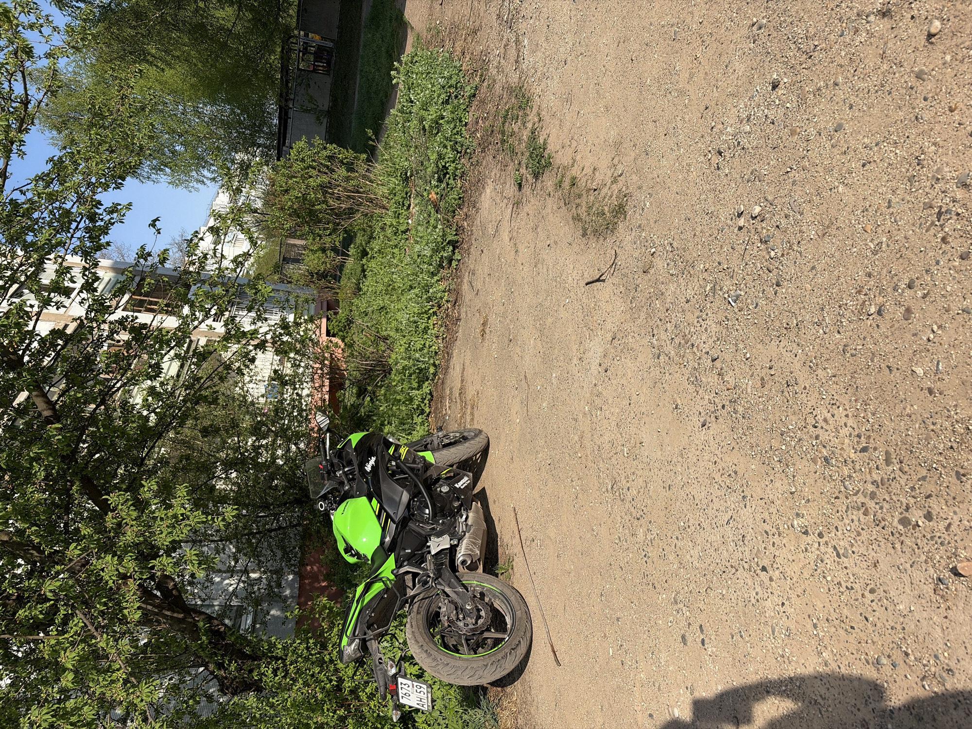 A green Kawasaki Ninja motorcycle is parked on a dirt road in Chaikovsky, Russia. The motorcycle is facing left. There is greenery and a building in the background. The license plate reads 7675 AH 59.