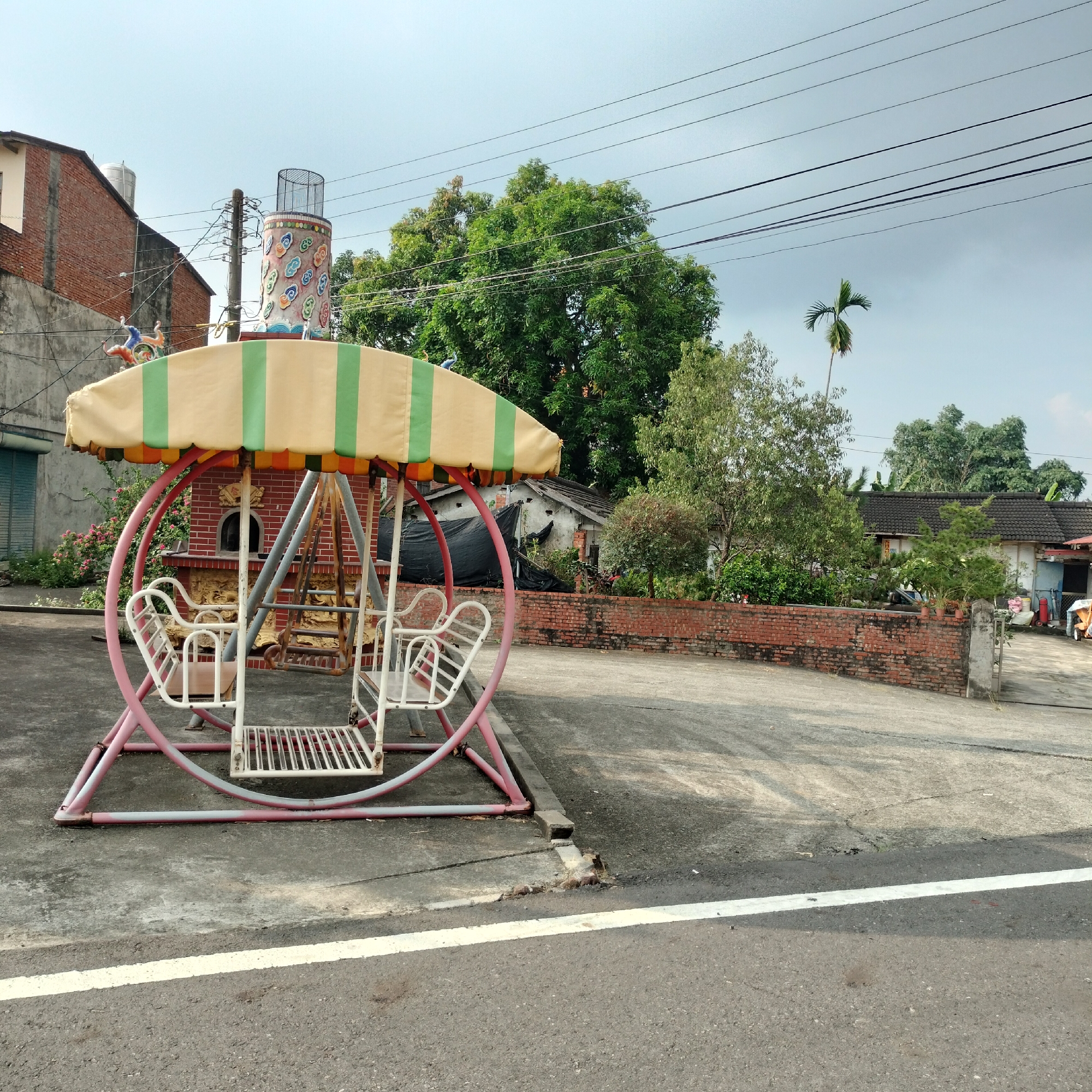 In Zhushan Township, Taiwan, a pink and white swing set with a striped yellow and green canopy stands on a concrete surface. The swing set features four seats, two facing each other, and is surrounded by a circular pink metal frame. Behind the swing set, a red brick building with an arched doorway is visible. To the left of the swing set, a two-story brick building with a light-colored facade and a water tank on the roof is present. A utility pole with numerous wires is situated between the two buildings. The background is filled with lush green trees and a cloudy sky. The scene is captured from a low angle, showing the swing set prominently in the foreground.