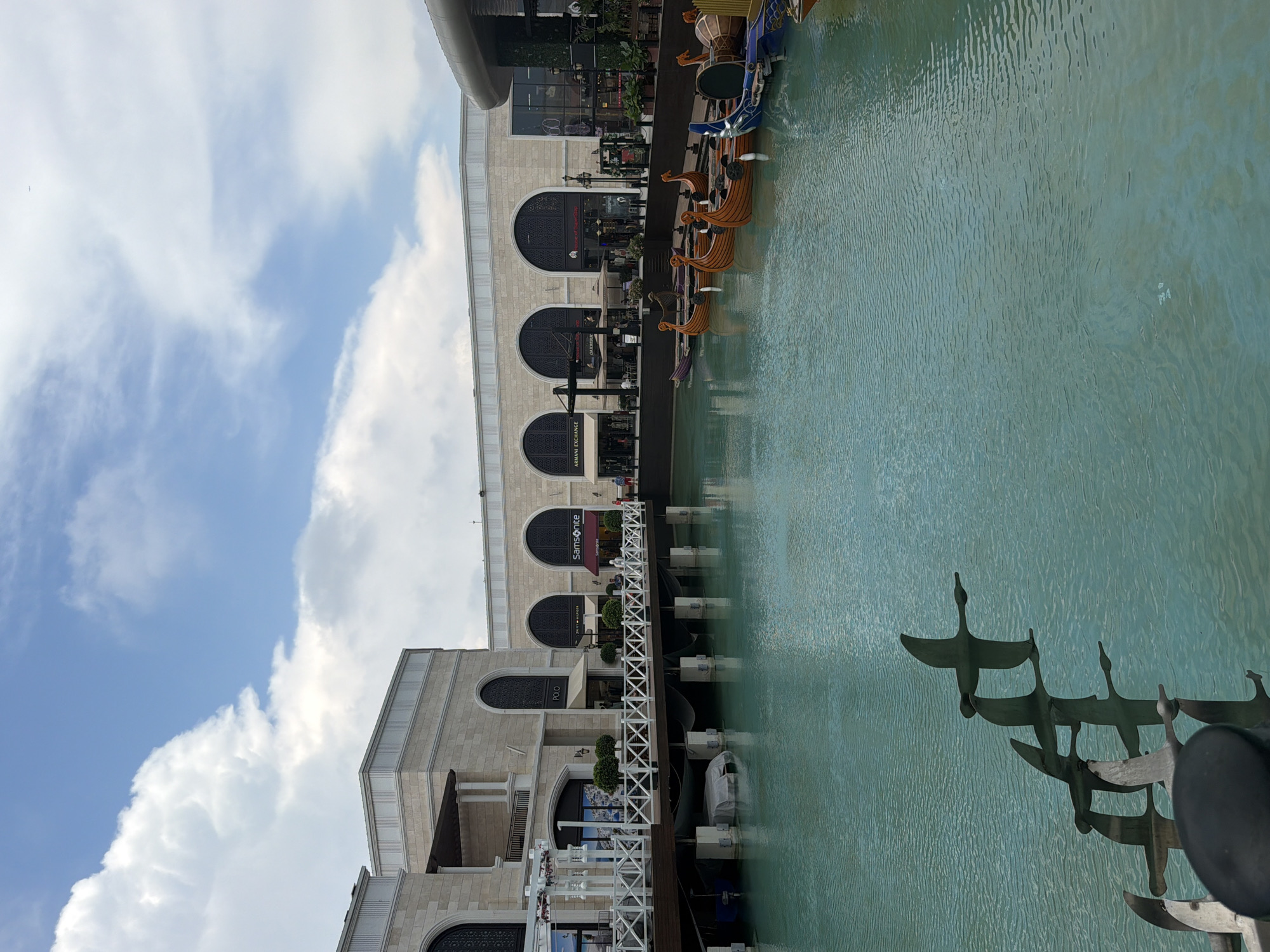 The image depicts a tranquil canal scene in Serik, Turkey, specifically within the Ligature Mall, a prominent shopping and entertainment destination. A bright blue sky with scattered white clouds forms the upper portion of the frame, contrasting with the light beige stone architecture of the mall buildings. The buildings feature large arched windows and colonnades, suggesting a Mediterranean or classical architectural style.
Along the canal's edge, a white wooden boardwalk with railings provides pedestrian access to the shops. Several storefronts are visible, including "Polo," "Samsonite," and "Armani Exchange," indicating a retail environment. Small, decorative green shrubs are interspersed along the boardwalk.
The canal itself is filled with a vibrant, turquoise-colored water, calm with gentle ripples. Moored along the far side of the canal are several decorative wooden boats, designed in a style reminiscent of Viking longships, with ornate prows and sterns.
In the foreground, a metallic sculpture of three birds in flight is partially submerged in the water, their reflections visible on the surface. The overall impression is one of a modern, upscale leisure and shopping complex designed to evoke a picturesque waterside setting.