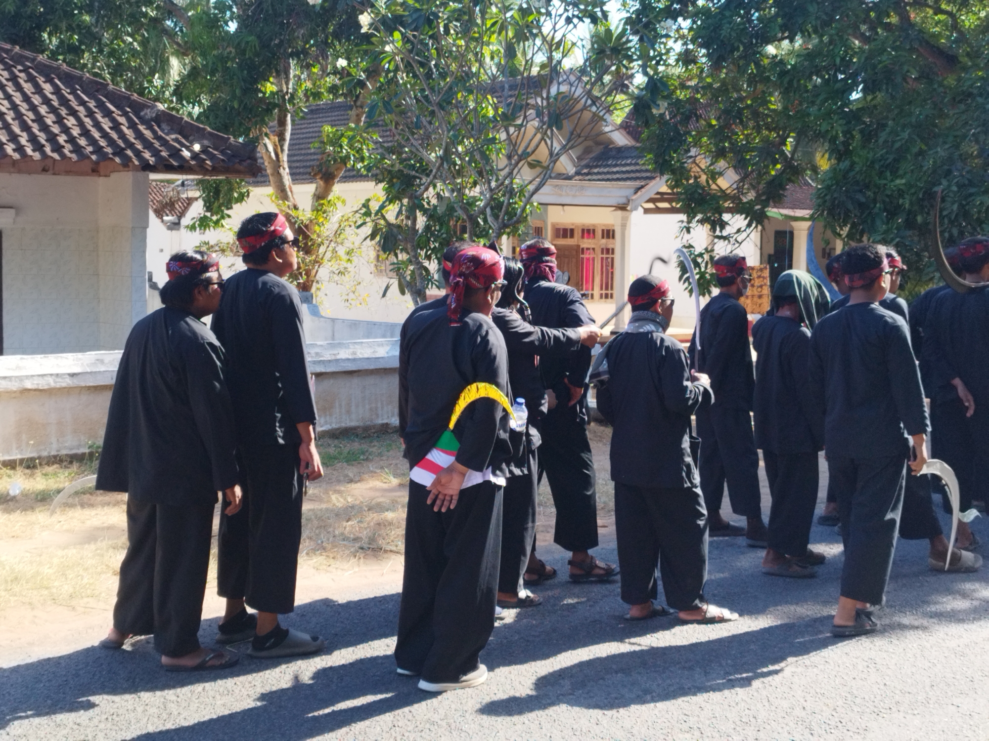 In Andulang, Indonesia, a group of men dressed in traditional black clothing and red headbands stand on an asphalt surface outdoors. The attire includes loose-fitting pants and long-sleeved shirts. Several individuals are holding curved, blade-like objects, possibly ceremonial weapons. One man in the foreground has a yellow and white sash draped across his back. The background features a white building with a tiled roof and lush green trees. The scene appears to be a cultural event or procession.