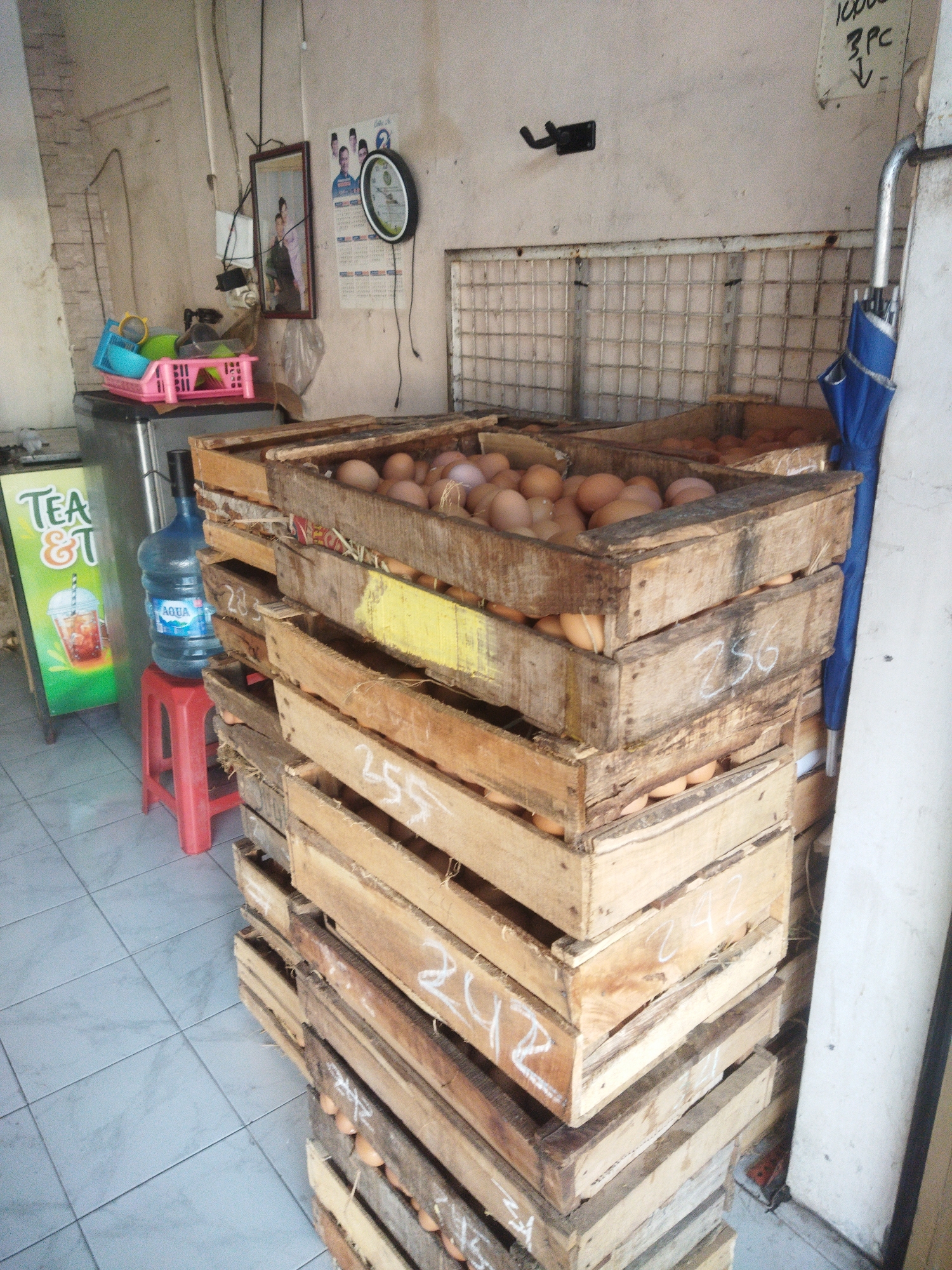 This image depicts an interior scene, likely a small shop or market stall, in Sukoharjo, Indonesia. The foreground is dominated by a stack of wooden crates filled with brown eggs. The crates are piled high, with some showing handwritten numbers in white chalk, such as "256" and "242". The eggs appear to be fresh and are roughly uniform in size and color.

To the left of the crates, a large blue jug of Aqua bottled water rests on a small red plastic stool. Adjacent to the water jug is a green and white sign advertising "TEA &", with an image of an iced tea drink. Behind the stool and water jug, a silver refrigerator is partially visible.

On the wall in the background, a framed photograph of two people is hanging, and next to it, a circular clock with a black frame and white face displays the time. A blue umbrella is leaning against a white pillar on the right side of the image. The walls are textured, and the floor is tiled with light blue square tiles. The overall impression is of a humble, functional retail space.