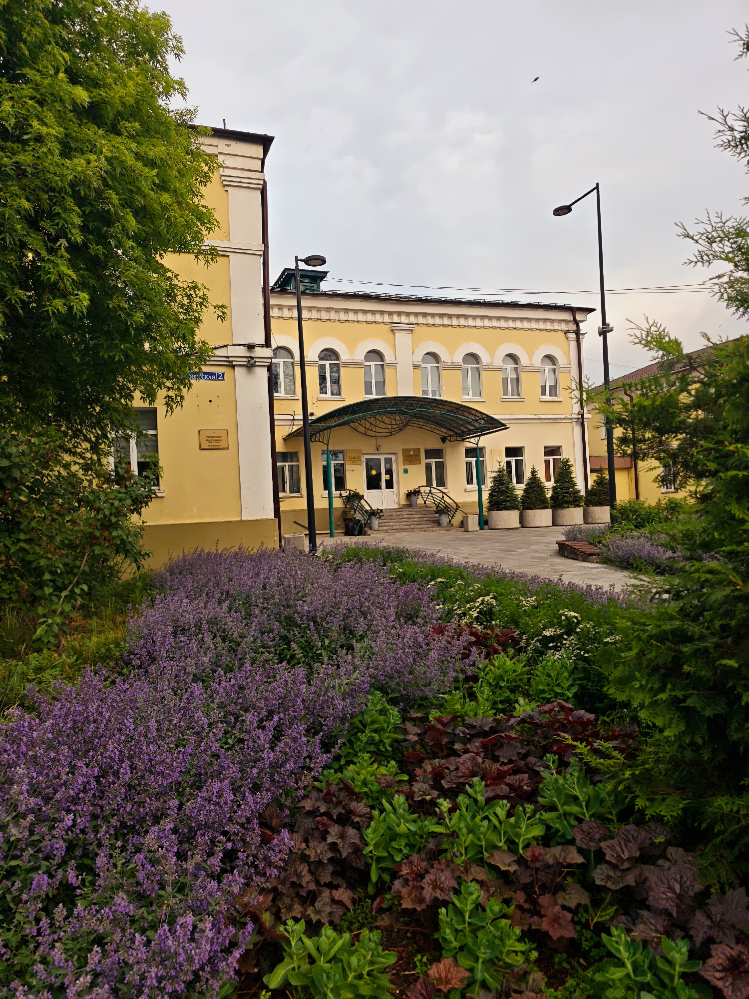 In the city of Unknown, Unknown, this image captures a building with a yellow facade and a dark brown roof. The building features a series of arched windows on its upper level and a canopy-covered entrance with stairs leading up to it. In front of the building, a pathway is flanked by a variety of plants, including a dense bed of purple flowers in the foreground and darker, broad-leafed plants. To the left, a large green tree with textured leaves partially obscures the building. Several potted evergreen trees are positioned along the pathway. A street lamp stands on the right side of the image, and a bird is visible in the overcast sky. The overall scene suggests a well-maintained exterior of a public or institutional building.