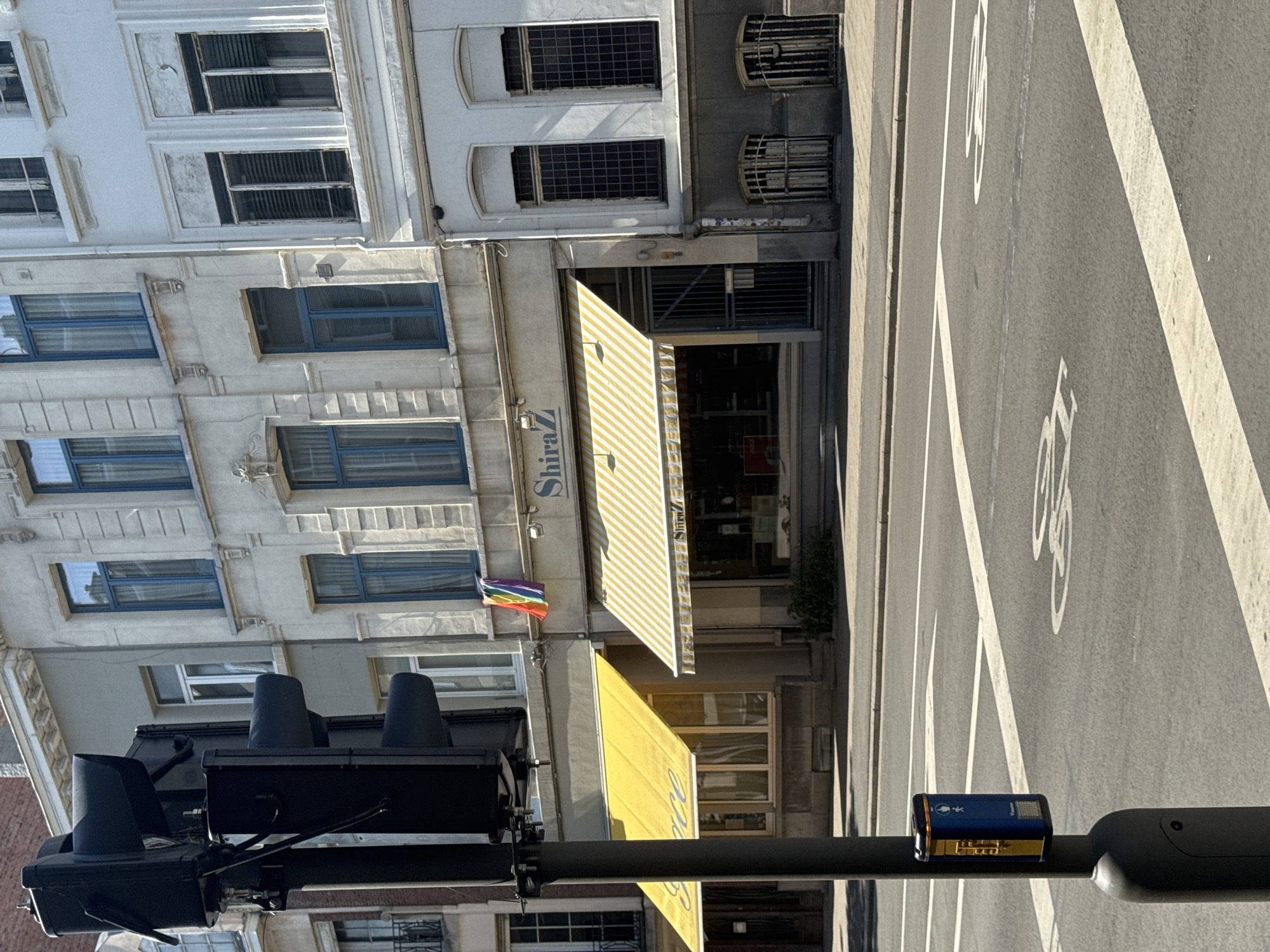 The image shows a street scene in the city of Antwerp, Belgium. In the foreground, a black traffic light pole is visible on the left side of the frame. Attached to the pole is a blue and yellow pedestrian crossing signal box with an illuminated pedestrian symbol. The street in front of the buildings has white lane markings and a bicycle symbol painted on the asphalt.
Behind the traffic light, a row of European-style buildings with multiple stories is visible. The buildings have light-colored facades, many with decorative architectural details around the windows. Several windows on the upper floors have blue frames.
In the middle ground, two storefronts with yellow awnings are prominent. The awning on the left is solid yellow, and the awning on the right has a yellow and white striped pattern. Above the right awning, a small rainbow flag is displayed, hanging from one of the upper windows. The name "Shiraz" is visible on the facade above the right storefront, with the "S" capitalized and a stylized Z.
The overall environment appears to be an urban setting on a sunny day, with shadows cast by the buildings and objects. No people are clearly visible in the image. The scene depicts a typical streetscape with pedestrian infrastructure and commercial establishments.
