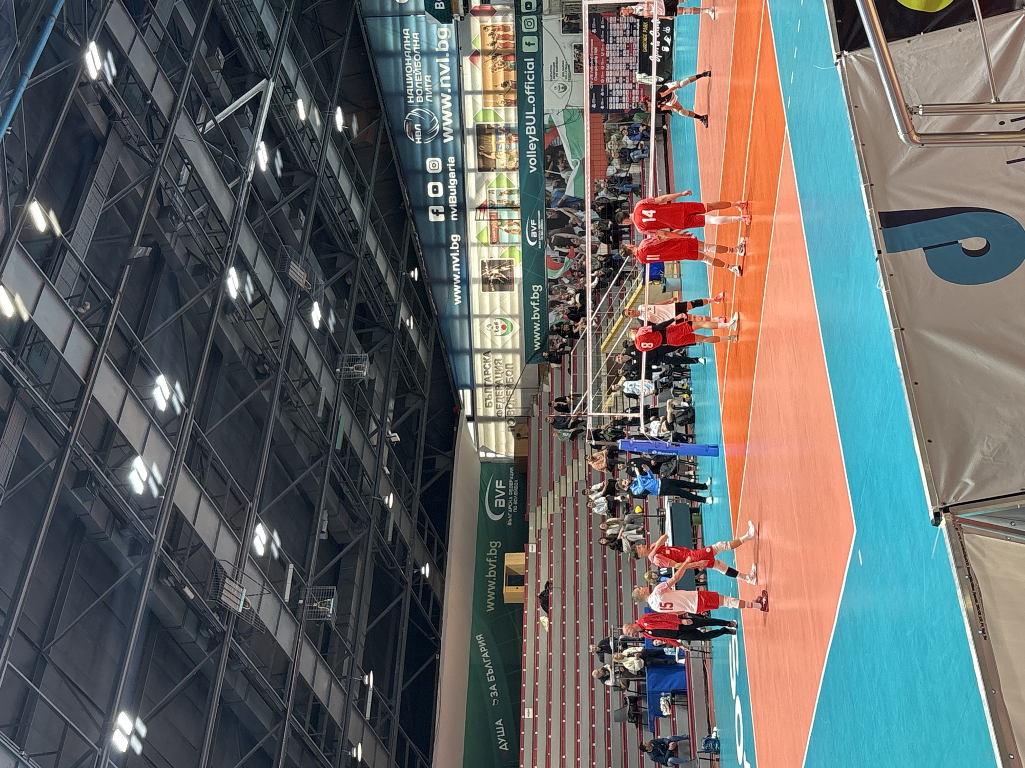 A volleyball match is in progress on a blue and orange court inside a large indoor arena. A team wearing red uniforms lines up near the net, while a player from the opposing team prepares to serve. Spectators fill the stands in the background. Banners with website addresses and social media icons are displayed around the arena. The location is an unidentified building or facility.