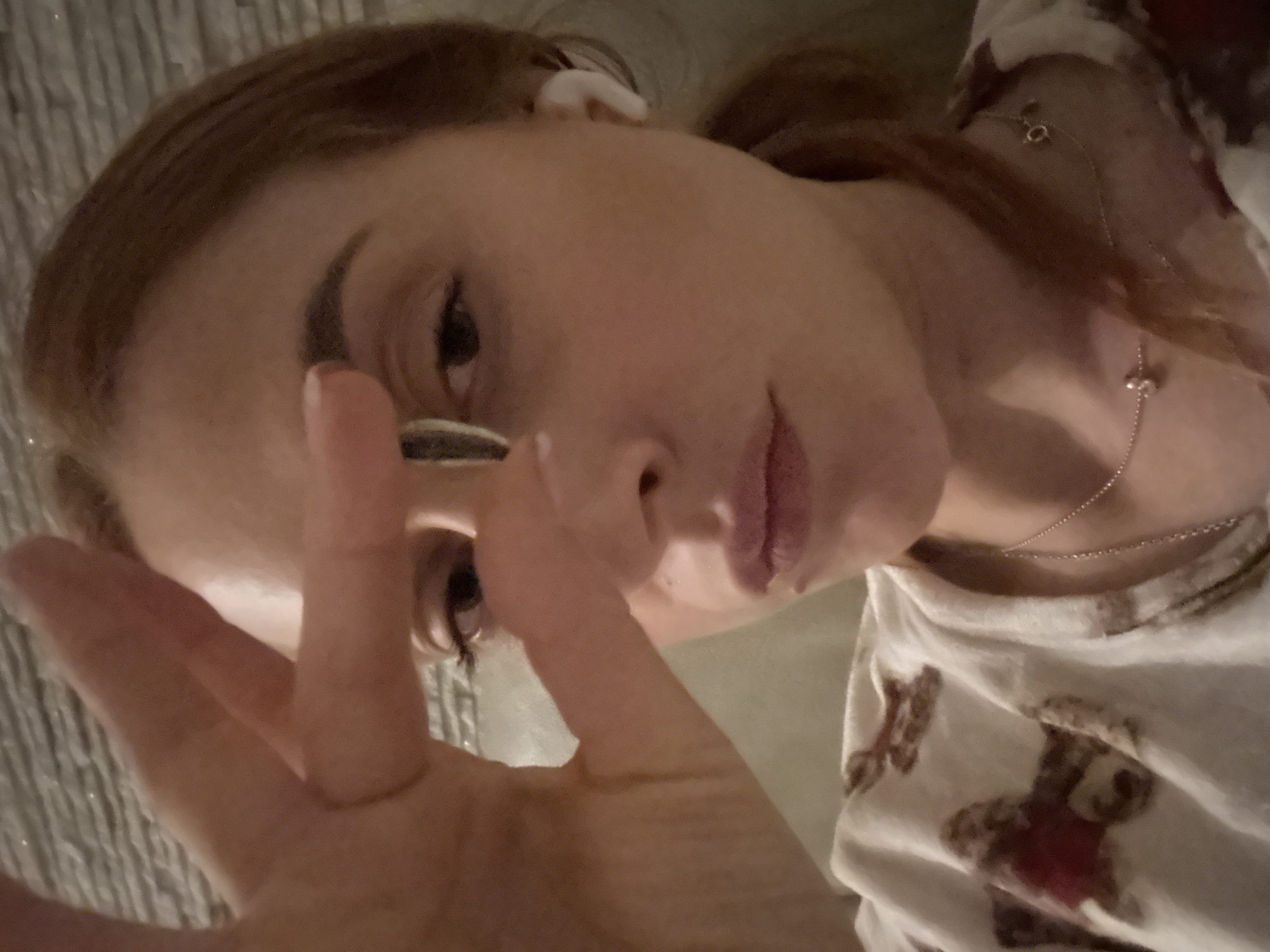 A selfie of a fair-skinned woman with reddish-brown hair is taken from a close-up, low-angle perspective. She wears a white shirt adorned with teddy bear motifs. A thin silver necklace graces her neck. The background is blurred, with a glimpse of what appears to be textured bedding. Her right hand is extended towards the camera, pinching a small, circular object between her fingers. In her right ear, an earbud is visible. The location in the city of Budva, Montenegro is not discernible from the visual information provided.