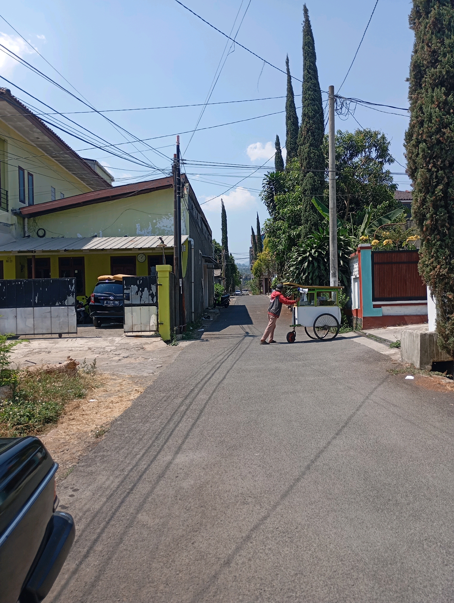 The image depicts a narrow, paved street in a residential area of Cimahi, Indonesia, under a clear blue sky. On the left, a two-story house with a yellowish-green facade and a dark tiled roof is visible, with a black metal gate and a yellow post. A black car with license plate "D 1191 FBG" is parked behind the gate, along with a motorcycle. Tall, slender cypress trees line the street, interspersed with other greenery and a building with a colorful facade on the right. In the middle of the street, a person wearing a red and gray jacket and light-colored pants is pushing a food cart with a white body and green accents. The cart has two large wheels and one smaller wheel at the front. Power lines crisscross above the street. The scene appears to be captured on a bright, sunny day, casting distinct shadows on the asphalt.