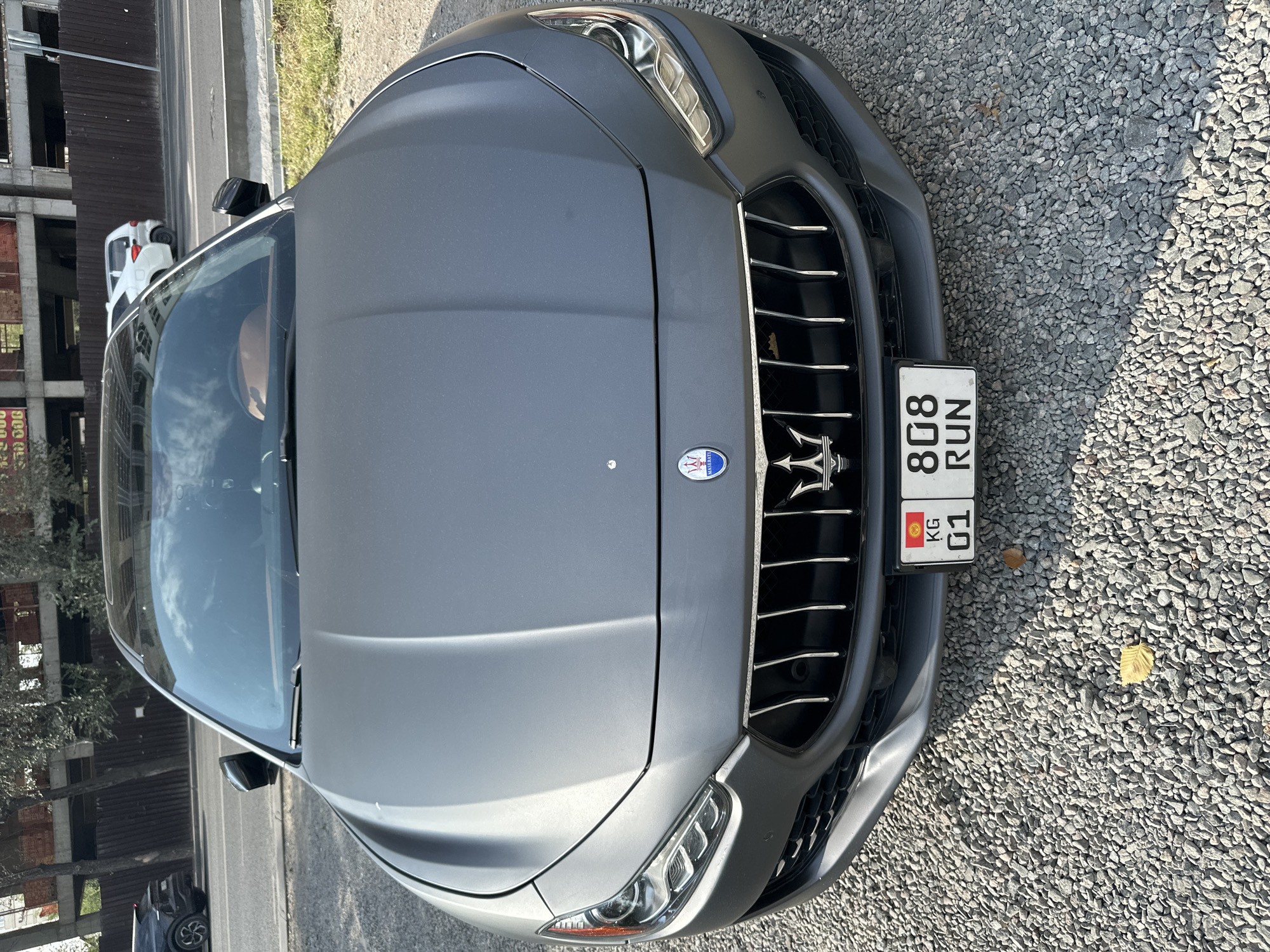 This is a front view, medium shot, eye-level photograph of a gray Maserati Ghibli parked on a gravel surface in Bishkek, Kyrgyzstan. The car's hood is prominent in the foreground, reflecting the sky. The Maserati emblem and trident are visible on the grille. A license plate with Kyrgyz insignia and the characters "KG 01 808 RUN" is mounted below the grille. In the background, a street runs alongside the car, with a dark-colored SUV and a white hatchback parked on the side of the road. Construction fencing and buildings are visible beyond the road. The environment appears to be outdoors on an overcast day, with some scattered leaves on the gravel.