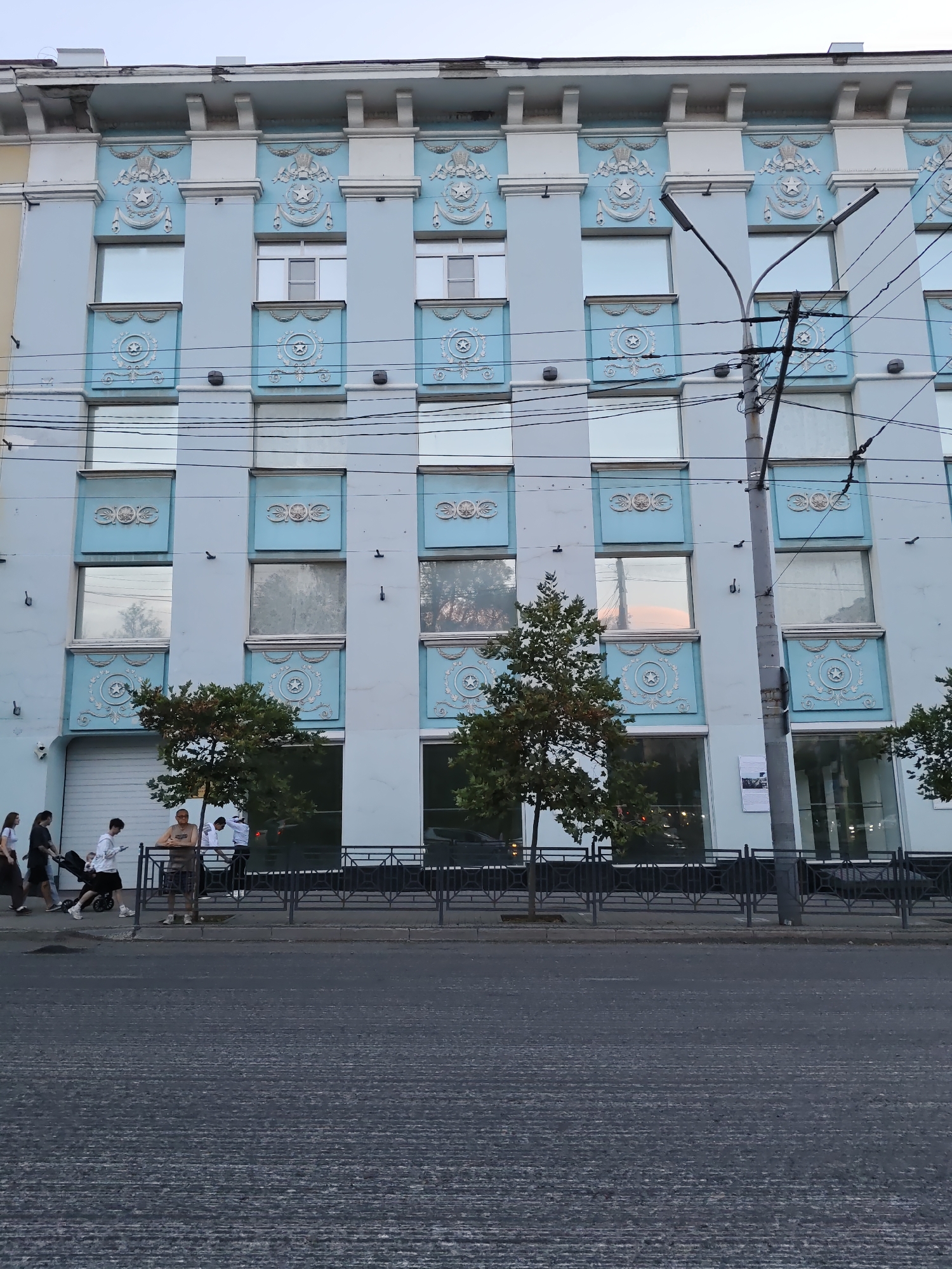 The image captures the facade of a light blue, multi-story building on a street in Rostov-on-Don, Russia. The building features a symmetrical design with rows of rectangular windows framed by ornate blue decorative elements that contain star motifs. Above the windows, there are elaborate white plaster decorations resembling crowns and wings, set against a white frieze. A lamppost with multiple arms and numerous power lines are visible in front of the building.
At the bottom of the building, a row of storefront windows is visible, some reflecting the sky and trees. A dark metal fence runs along the sidewalk in front of the storefronts. On the sidewalk, several individuals are present. On the left, a group of people are walking, including a person pushing a stroller. Further to the right, a man stands leaning against the fence, and another man in a white shirt is also near the fence. Trees are planted along the sidewalk. The street in the foreground is paved with asphalt, showing textured lines. The sky is overcast.