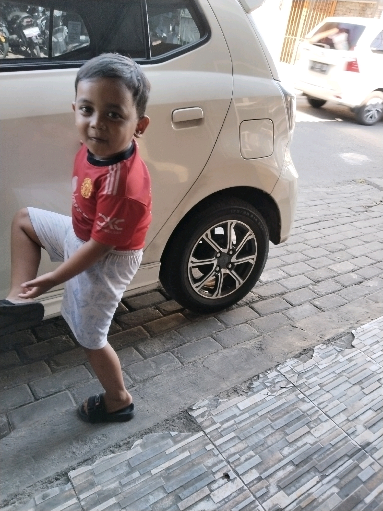 A young boy is captured in a full-body shot, posed playfully with one leg raised against the side of a light-colored car. He is wearing a red t-shirt with white stripes on the sleeves and a white, patterned pair of shorts. The boy's hair is dark and short, and he is looking directly at the camera with a slight smile. He is wearing black sandals. The car, likely a hatchback, is light beige and parked on a paved surface that appears to be a sidewalk or driveway made of interlocking rectangular stones. In the background, another light-colored car is visible, seemingly in motion, and a building with a striped metal fence can be seen. The setting suggests a residential or public outdoor space in a city. The scene is illuminated by natural daylight.
