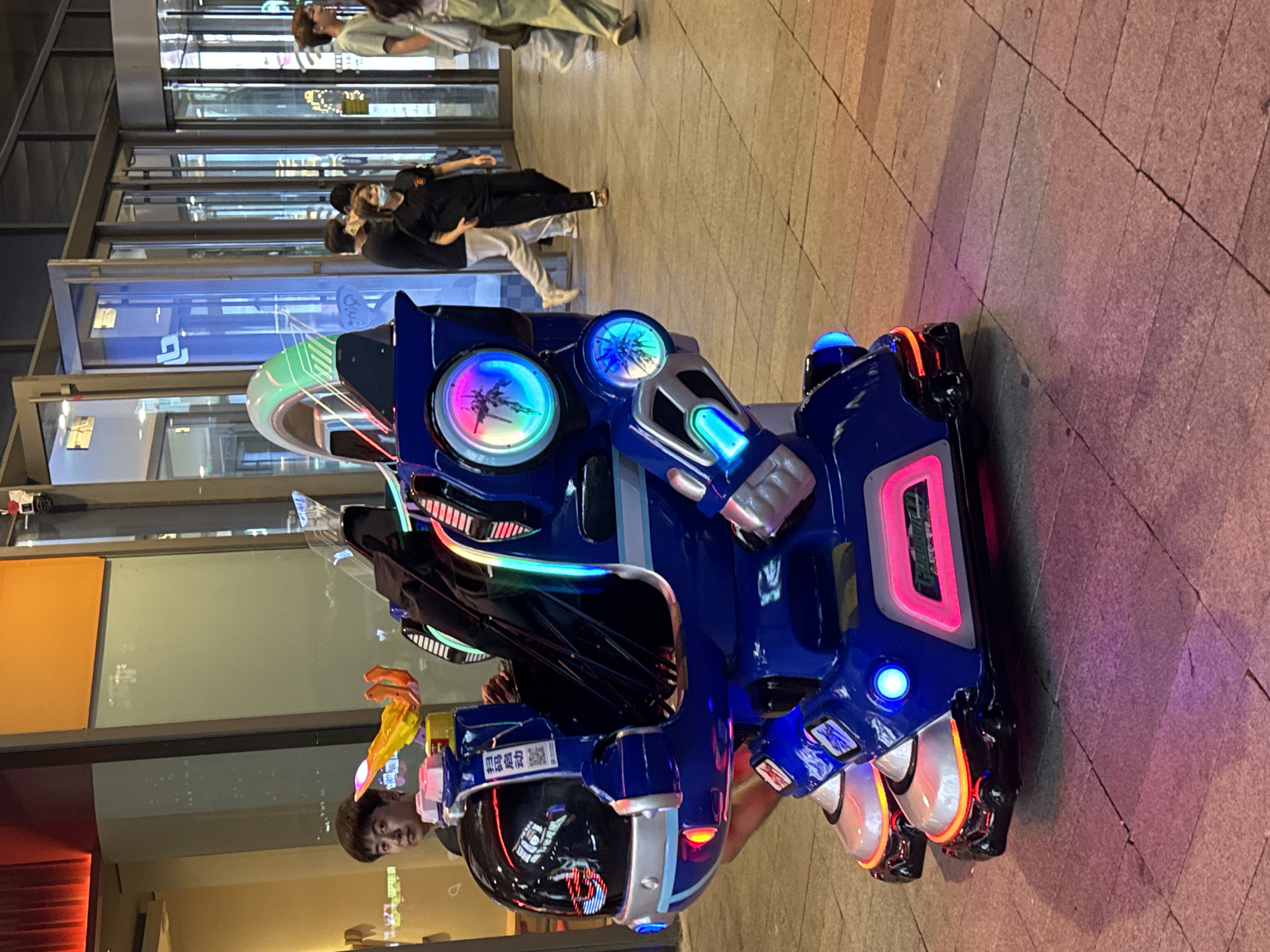 In an urban outdoor setting, possibly a pedestrian walkway or plaza in Shenyang City, China, a vibrantly lit blue and silver robotic ride-on toy for children is the central focus. The toy, designed to resemble a mecha, is illuminated with various colored LED lights, including pink, blue, green, and yellow, creating a futuristic and eye-catching display. The toy is parked on a textured stone-tiled ground, with some tiles appearing cracked and worn.
To the left of the toy, a young Asian man with short dark hair is seated, partially obscured by the robotic structure. He is looking towards the camera with a neutral expression. He appears to be an attendant or supervisor of the ride.
In the background, a row of glass-fronted establishments suggests a commercial area or shopping mall. Several people are visible walking by, some dressed casually. One woman in a black t-shirt and dark pants is walking past the robotic toy, while another person in a light green t-shirt is further in the background. The lighting in the background is a mix of interior lights from the establishments and ambient evening light, casting reflections on the glass surfaces. The overall impression is of a lively public space at dusk or night, with the illuminated toy serving as an attraction.