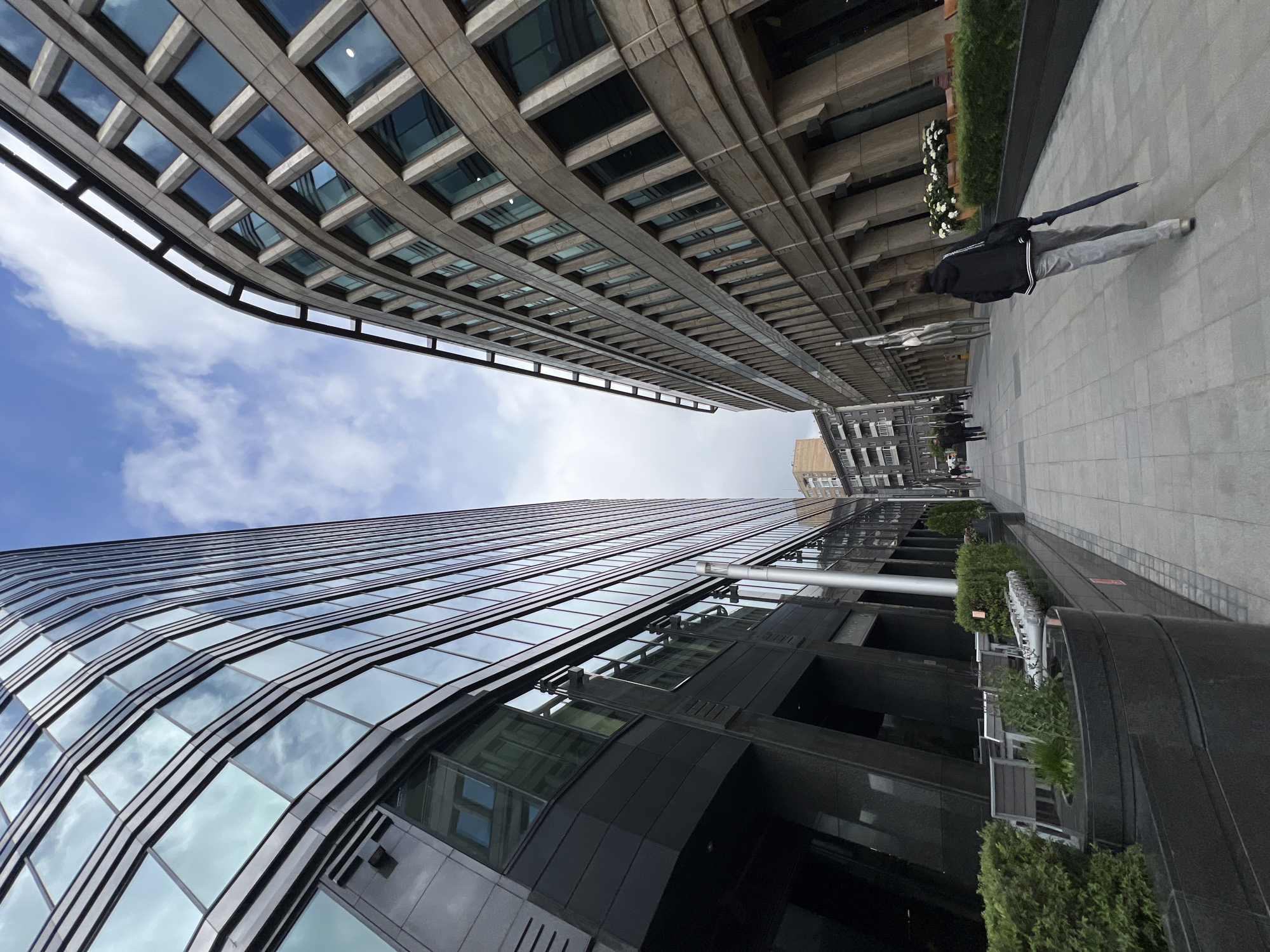 The image captures a street view in the business district of Moscow, Russia, characterized by towering modern architecture. Two high-rise buildings dominate the frame, one with a curved, glass-facade reflecting the sky, and the other with a more traditional concrete design featuring rows of rectangular windows. The perspective is from a low angle, emphasizing the height of the structures. On the right side of the image, a sidewalk runs alongside the building, with greenery planted along the edge. Several people are walking along the sidewalk in the distance, and one person is closer to the camera. The sky is visible between the buildings, showing scattered clouds. The photograph appears to have been taken during daylight hours.
