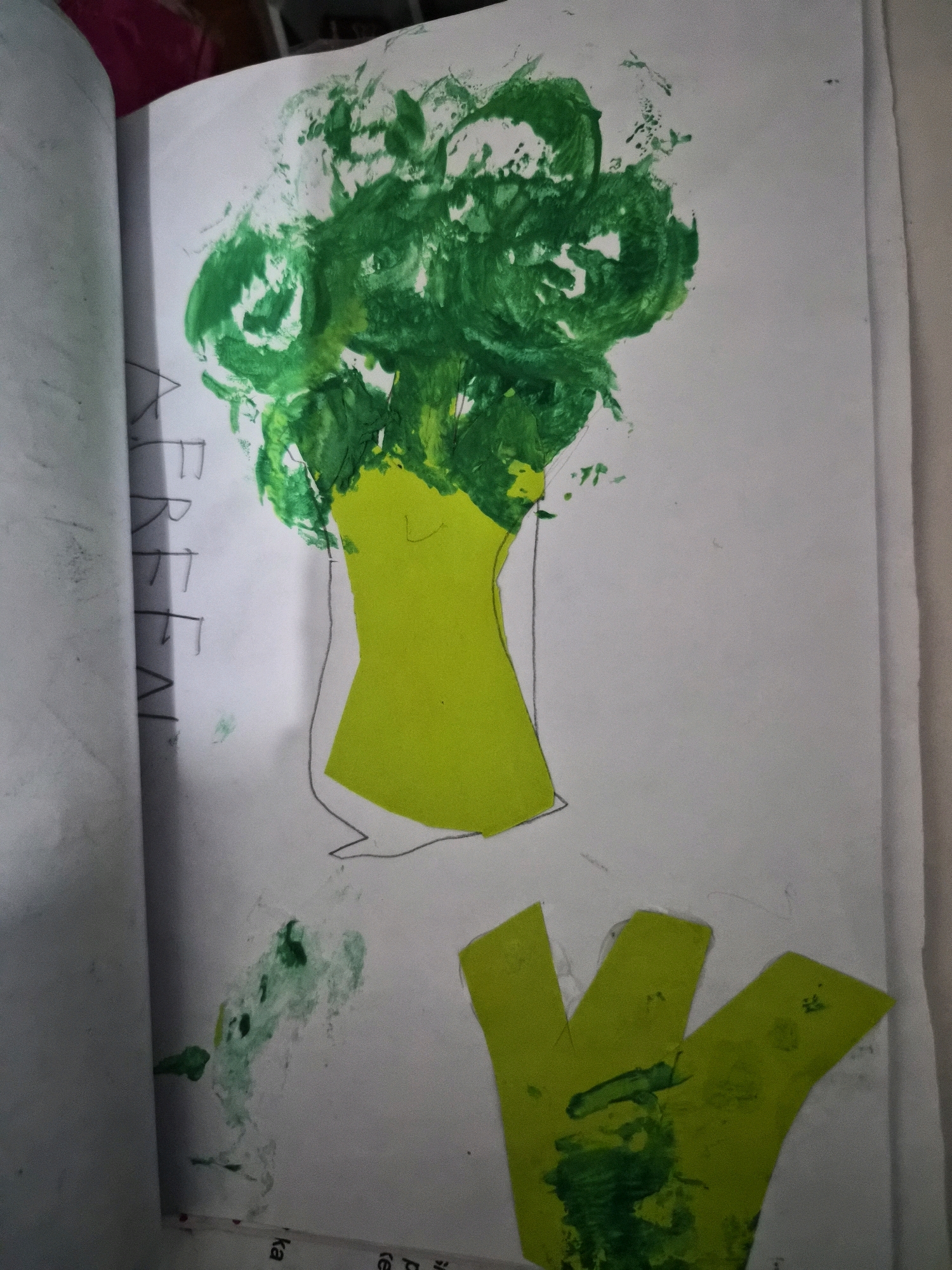 The image shows a child's drawing of a broccoli, created using paint and paper cutouts. The broccoli's florets are depicted with green paint, while the stem is a green paper cutout. Below the broccoli, there are three more green paper cutouts arranged in a fan shape, which could represent leaves or additional broccoli florets. Some green paint splatters are visible on the page. The drawing is in an open sketchbook or notebook, with a white page on the right and a partially visible left page containing faint pencil markings. The overall impression is that of a creative art project by a child. The provided image does not contain any discernible elements or context that would allow for its location to be identified in the city of Balikpapan, Indonesia.