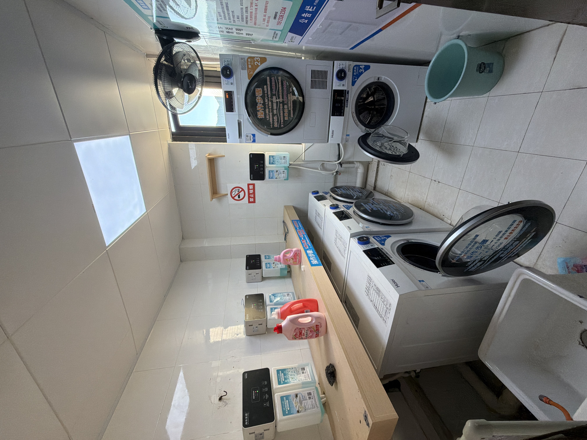 The image depicts a laundry room located in Jiangjin, China. The room is tiled with white ceramic tiles on the walls and floor. In the center of the image, a shelf made of light-colored wood holds several bottles of laundry detergent and two dispensing units. Below the shelf, several washing machines and dryers are stacked and side-by-side. A white Haier washing machine with its door open is visible on the right, and a dryer unit is stacked above it. To the left, another Haier washing machine is partially visible. In the foreground, a white sink is present. A teal laundry basket sits on the floor to the right of the washing machines. The room is illuminated by overhead lighting, and natural light streams in from a window in the background. A ceiling fan is mounted on the wall above the window. A "No Smoking" sign is displayed on the wall. The overall impression is of a clean and functional laundry facility. There are no people visible in the image.