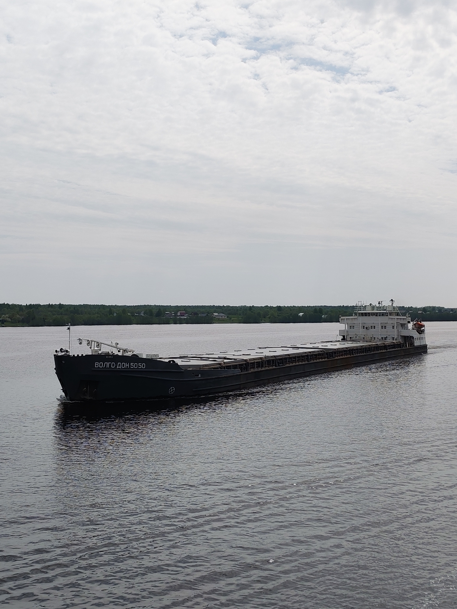 A wide-angle shot captures a cargo ship moving on a body of water, possibly a river or a lake. The ship, identified as "Волго-Дон 5050" is predominantly gray and black. A structure with multiple windows is visible at the back of the ship. The water is calm with ripples across its surface, ranging from a dark gray to a light gray. The opposite shore of the water body is lined with trees and a few visible structures. The sky is overcast. The location is in Раменье, Russia.