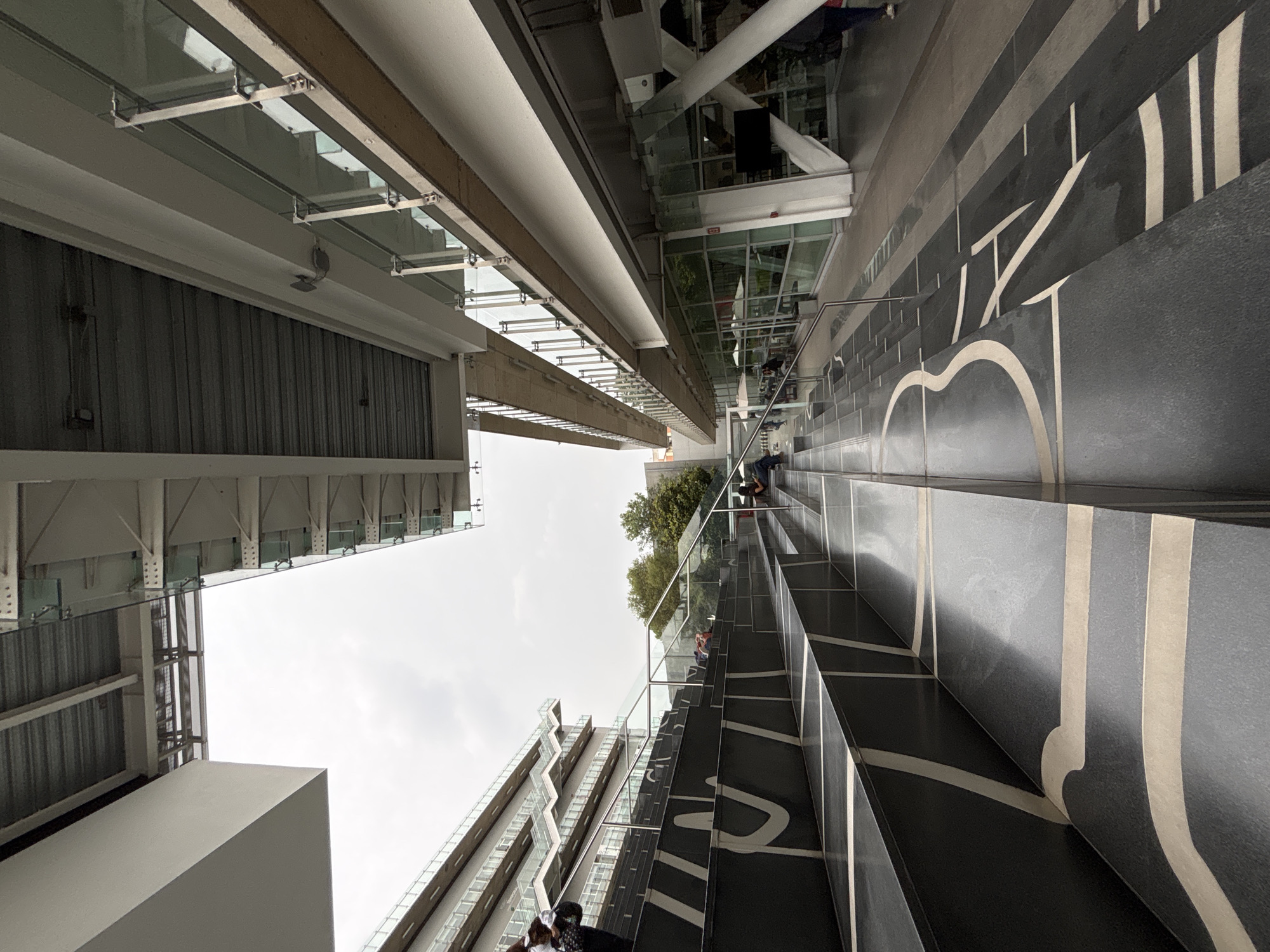 The image captures an upward view of the Papalote Museo del Niño in Mexico City, Mexico, taken from the base of the museum's prominent outdoor staircase. The staircase is constructed of dark-colored steps with bold, white calligraphic-style designs. People are visible on the staircase. The architecture surrounding the staircase is a mix of concrete, metal, and glass, giving a modern, urban feel. The sky is overcast. Buildings are visible on either side of the staircase. The shot seems to have been taken with the camera oriented vertically.
