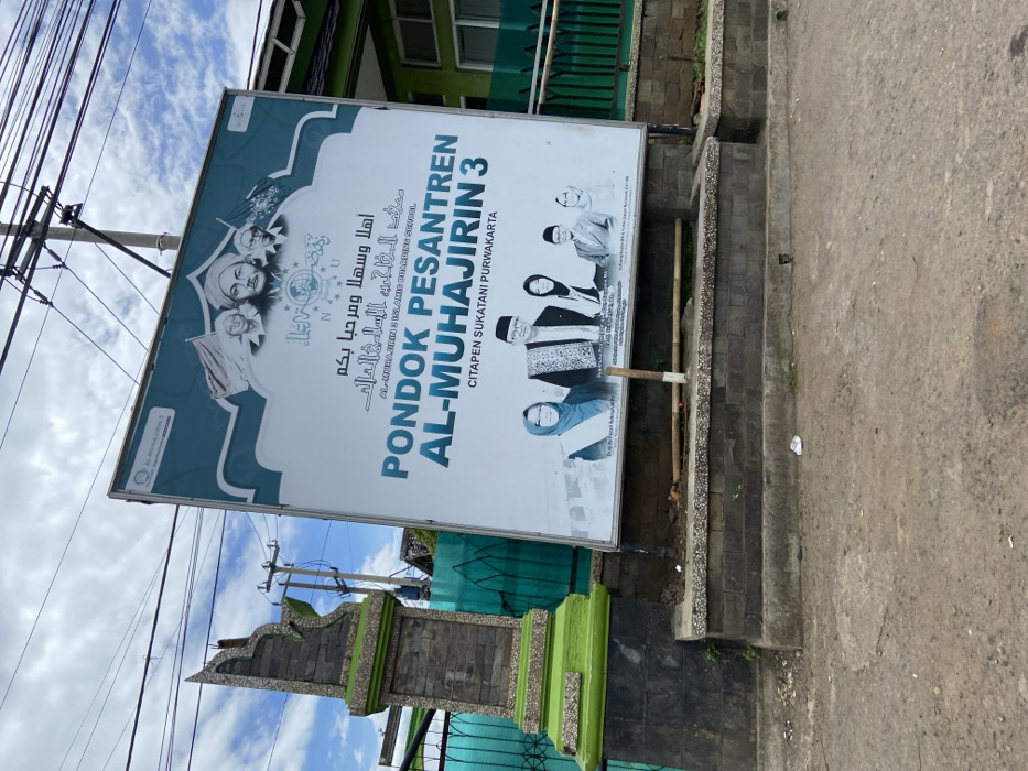 A large sign with the words "Pondok Pesantren Al-Muhajirin 3" in Arabic and Indonesian is mounted on a brick wall. The sign is located in front of a building in the city of Sukatani, Indonesia, at coordinates -6.596 latitude and 107.424 longitude. The sign has a white background and blue lettering. The sign features a picture of five people, likely administrators of the school, standing together in front of a green background. The people are wearing traditional Muslim clothing and smiling. The sign also features a decorative border and a small logo in the upper left corner. The sign is mounted on a brick wall that is partially covered in green paint. The sign is partially obscured by a small, green structure in the foreground. The building behind the sign has a green metal gate. The ground in front of the sign is made of concrete and gravel. There is a small amount of debris on the ground. The sky is overcast. The scene is daytime.