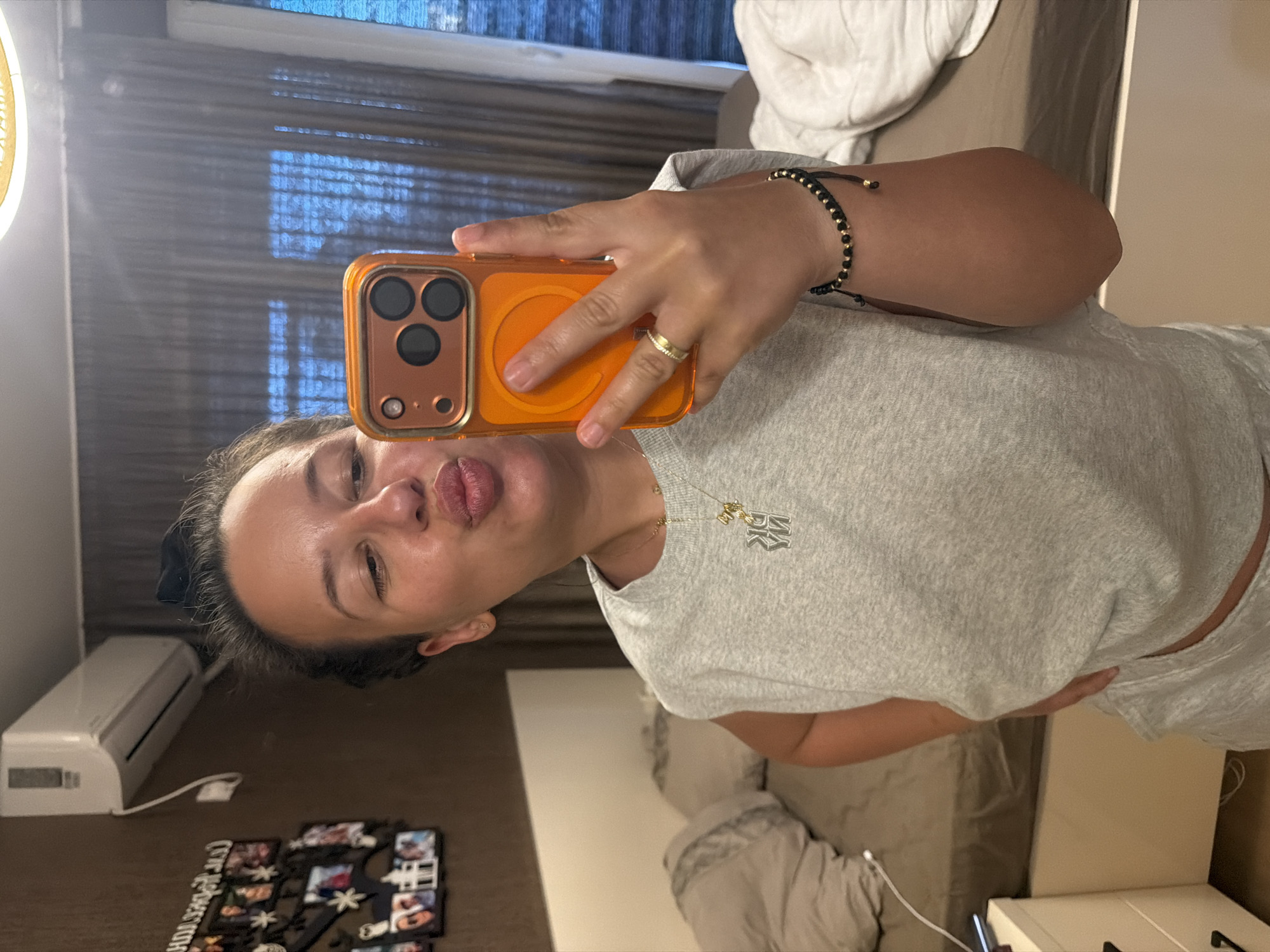 A woman is taking a mirror selfie in what appears to be a bedroom in Burgas, Bulgaria. She is wearing a grey sleeveless t-shirt and grey shorts, with a necklace and a bracelet. She is holding an orange phone in her right hand, taking the picture. The background shows a bed, a nightstand with a phone charger, a framed collage of photos on the wall, and an air conditioner unit. The room has a muted color palette with brown walls and beige curtains.