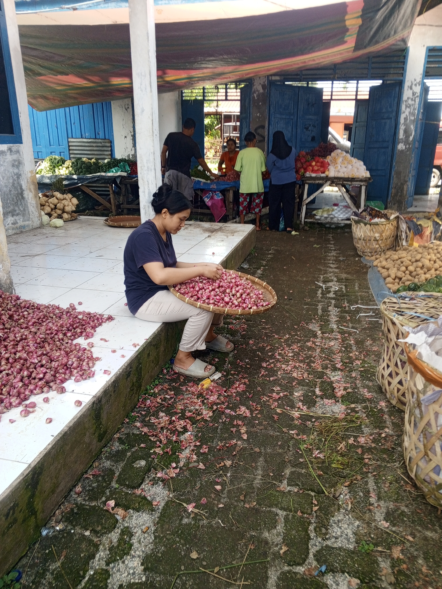 A woman is seated on a ledge at a market in Talun Kenas, Indonesia. She is wearing a dark blue t-shirt and beige pants, and is holding a large woven basket filled with small red onions. A large pile of red onions is spread out on the white tiled ground to her left. The ground around her is littered with onion skins and stems. In the background, other vendors are visible selling produce from stalls under a colorful tarp. The market appears to be an open-air structure with blue wooden doors.