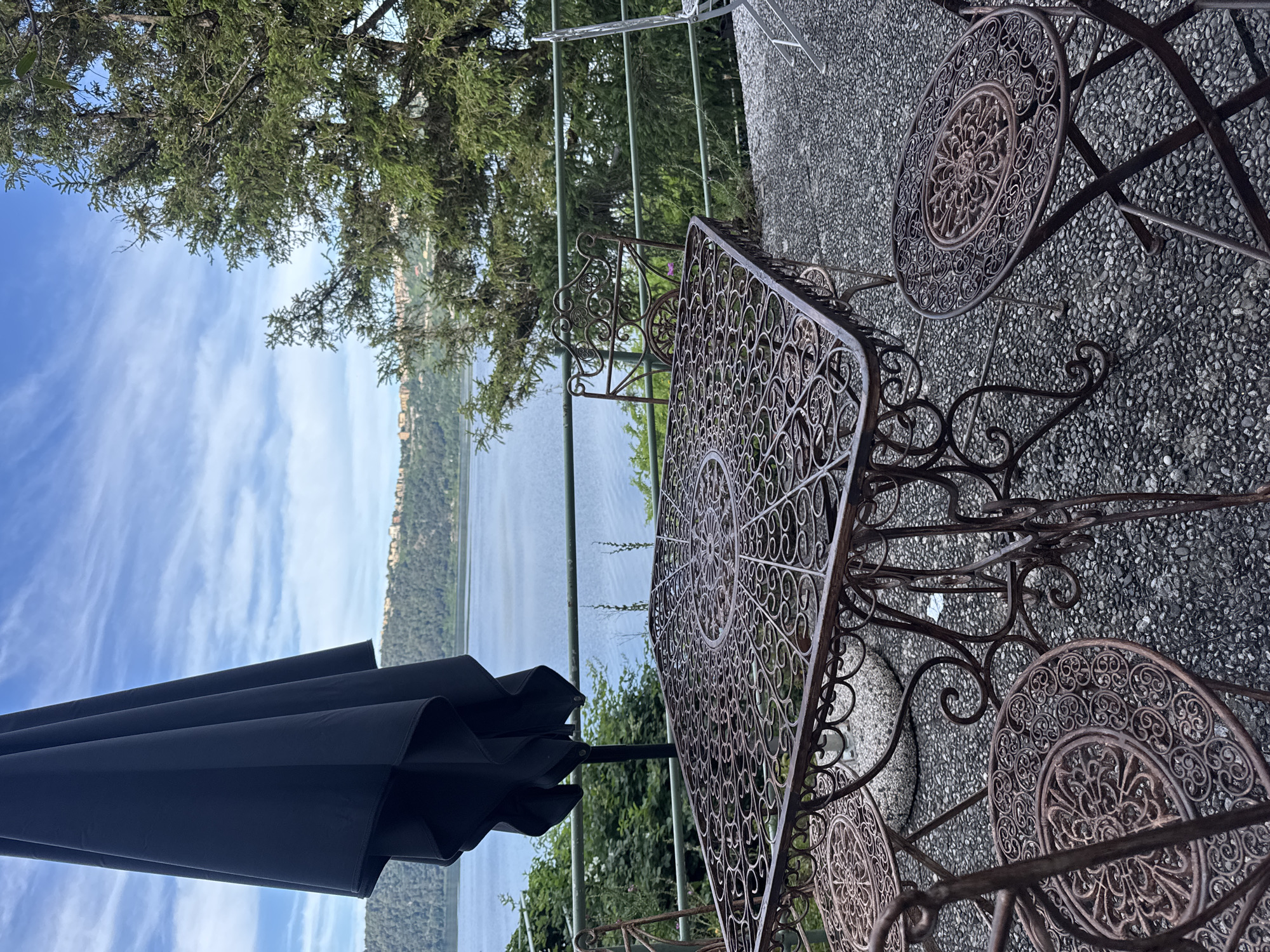 The image shows an outdoor seating area in Castel Gandolfo, Italy, overlooking a lake and distant hills. The seating set includes a square metal table with an ornate, openwork design and matching chairs, placed on a textured, pebbled surface. A black umbrella is partially visible, offering shade. A metal railing separates the seating area from the view of the lake. In the distance, a land with buildings are seen. Above, the sky is blue with scattered clouds, visible through the leaves of a tree.