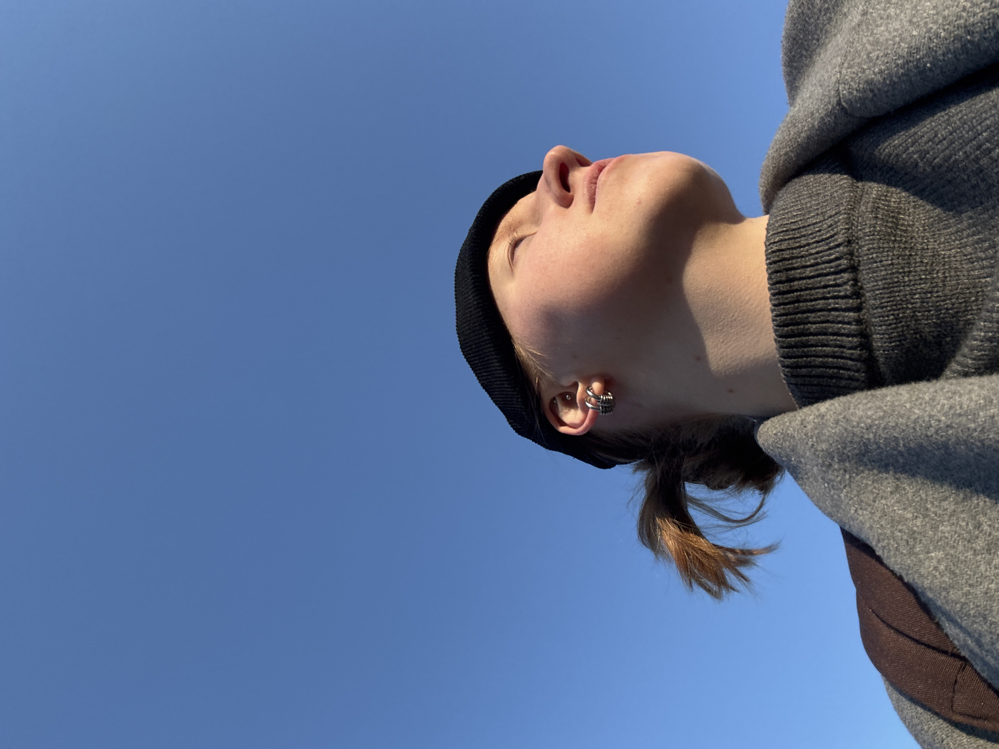 A low-angle shot depicts a person wearing a black beanie and a gray hooded jacket. The person is looking upwards towards a clear blue sky. A silver earring is visible in their ear. The person's hair is brown and pulled back into a low ponytail. The exact location in Perm, Russia, is not discernible from this image.