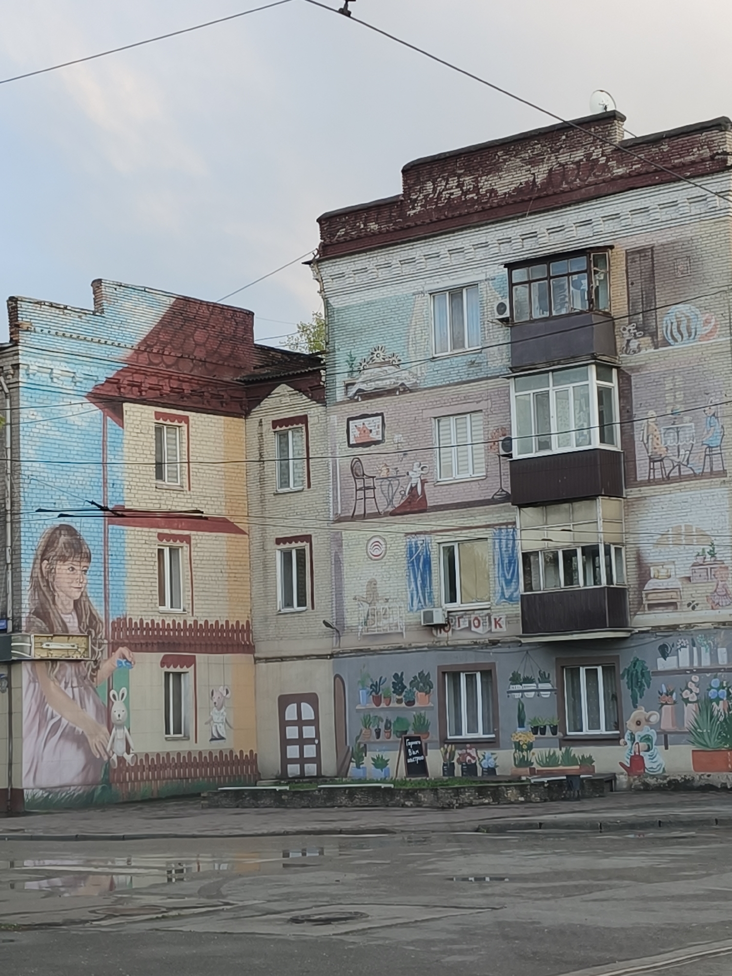 A multi-story brick residential building in Kryvyi Rih, Ukraine, is extensively covered in a vibrant, whimsical mural. The artwork transforms the facade into a detailed storybook-like scene, painted over the light-colored brickwork.
On the left side, a large mural of a young girl with long brown hair and glasses, wearing a light dress, is depicted, holding a small object. Below her, a white toy rabbit sits on a painted brown picket fence. This section's background is a light blue sky with white clouds. Continuing across the building's various sections, the mural portrays interior scenes featuring anthropomorphic mouse characters engaged in domestic activities, such as sitting at tables with furniture. A prominent feature is a section with numerous potted plants displayed on shelves, resembling a flower shop, where a mouse is depicted watering a plant. A small, painted brown door is visible at ground level. Painted red-tiled roofs are incorporated into the mural design on architectural extensions of the building.
The building's real windows and a multi-level, dark brown enclosed balcony are seamlessly integrated into the painted landscape. The asphalt ground in the foreground is wet with puddles, suggesting recent rainfall. The sky above is partly cloudy, indicating daytime.
Visible Cyrillic text includes "ПОГЛОК" on the building wall below the middle balcony. A black easel-style sign near the plant section reads "Гарного Вам настрою" (Have a good mood) in Ukrainian.