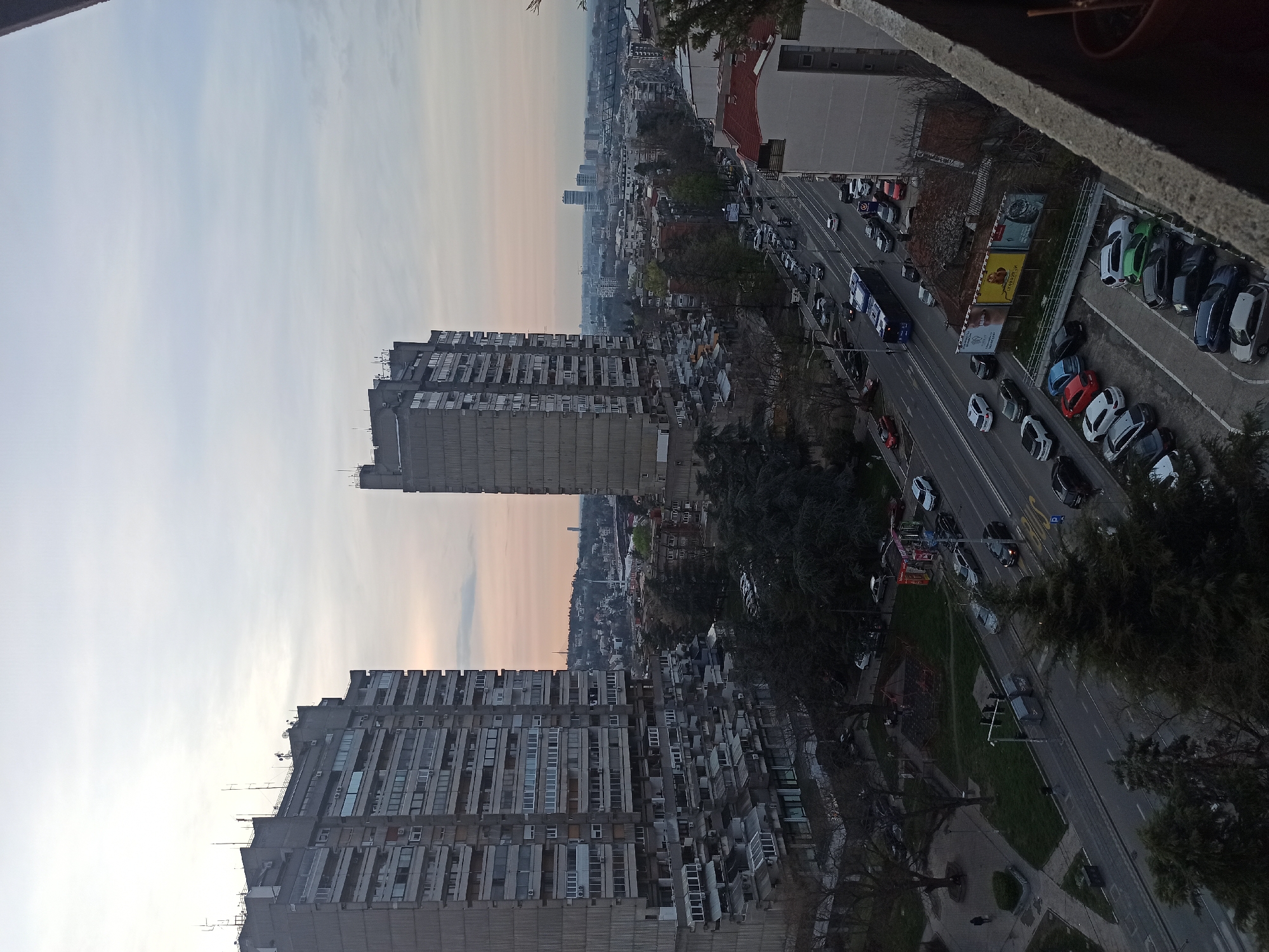 The image depicts an elevated view in the Vozdovac Urban Municipality, Serbia. In the foreground, two tall, concrete apartment buildings with numerous windows and balconies dominate the scene. A road runs parallel to the buildings, filled with cars indicating traffic. Adjacent to the road, a parking area contains a variety of parked cars. Billboards are visible along the road. Beyond the apartment buildings, the cityscape extends into the distance with a mixture of residential and commercial structures. The sky is overcast with hues of gray and beige. Trees are interspersed throughout the urban landscape.