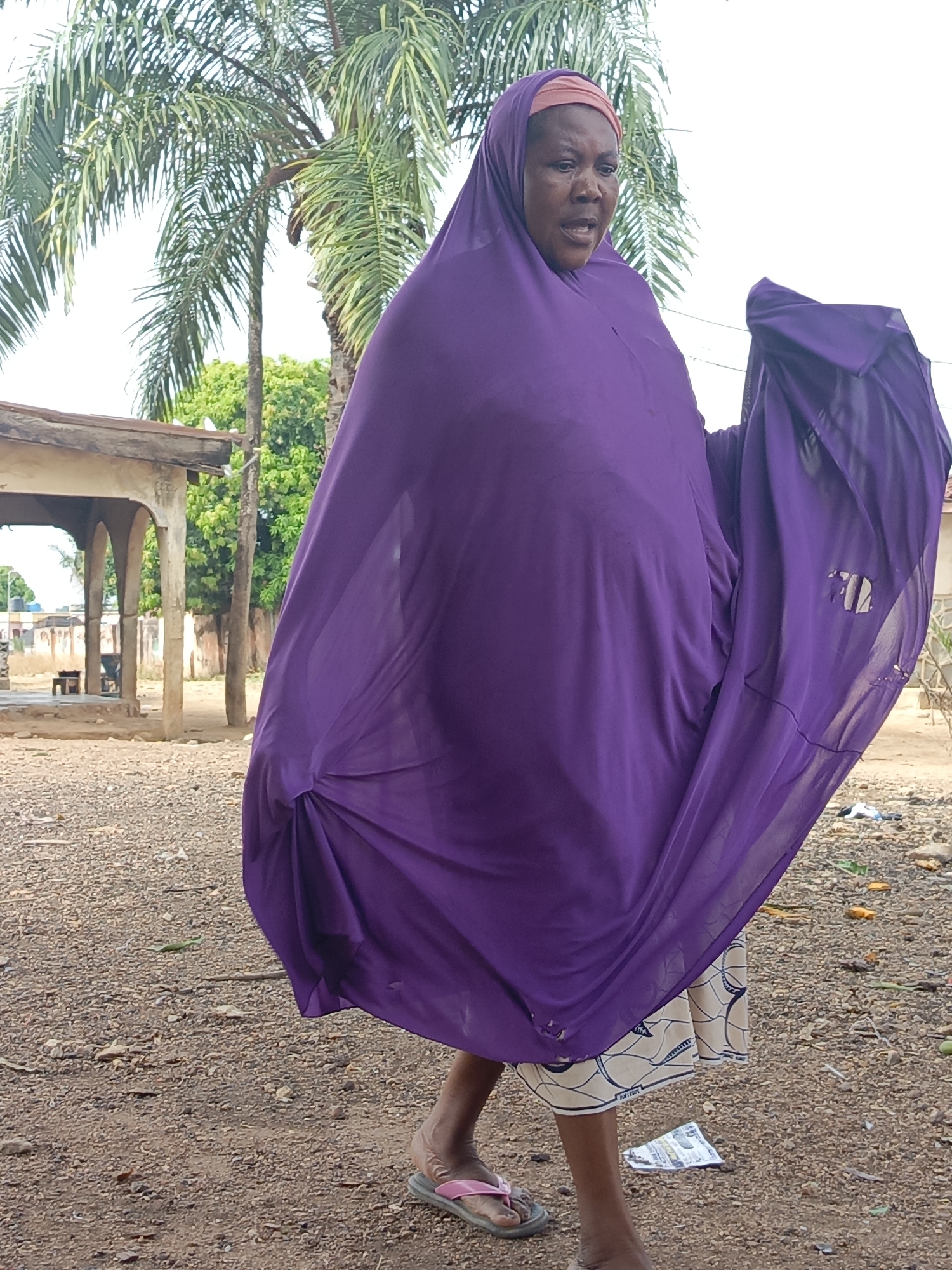 An adult woman is centered in an outdoor scene, wearing a long, flowing purple garment that covers most of her body and head. She wears a peach-colored head covering beneath the purple garment. The woman’s facial expression appears to be one of distress. She wears sandals on her feet.
The ground is covered in dirt and gravel, with scattered trash and debris. A building with a covered walkway and pillars is visible in the background to the left of the woman. A tall palm tree is also visible behind her.
Based on the information available, this scene is taking place in Akwanga, Nigeria.