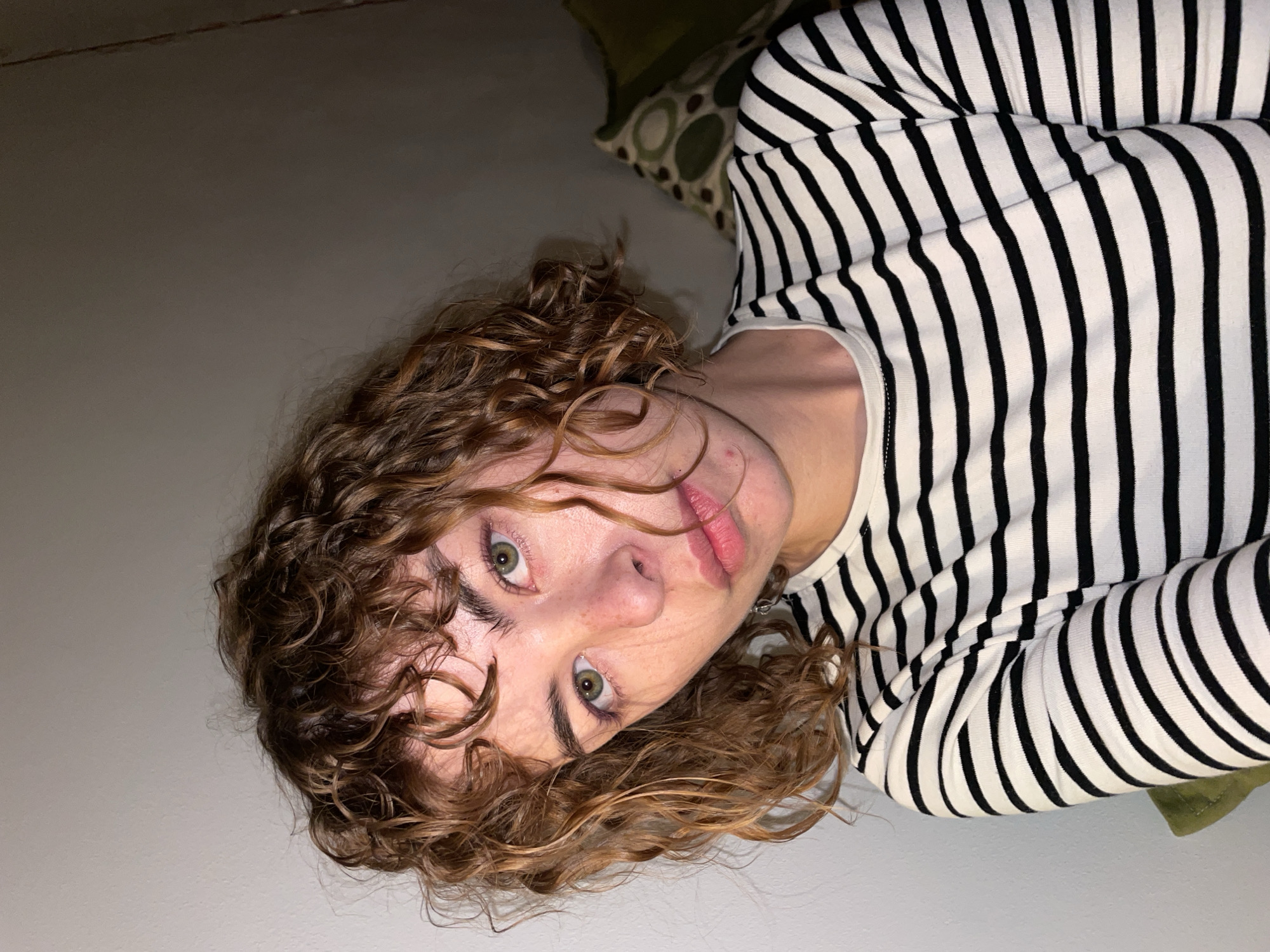 The image is a close-up shot of a person with curly, brown hair wearing a black and white striped shirt. The person has fair skin and green eyes. The location is likely indoors, with a plain wall in the background on one side and a patterned pillow on the other. There is no indication of Boreti, Montenegro.